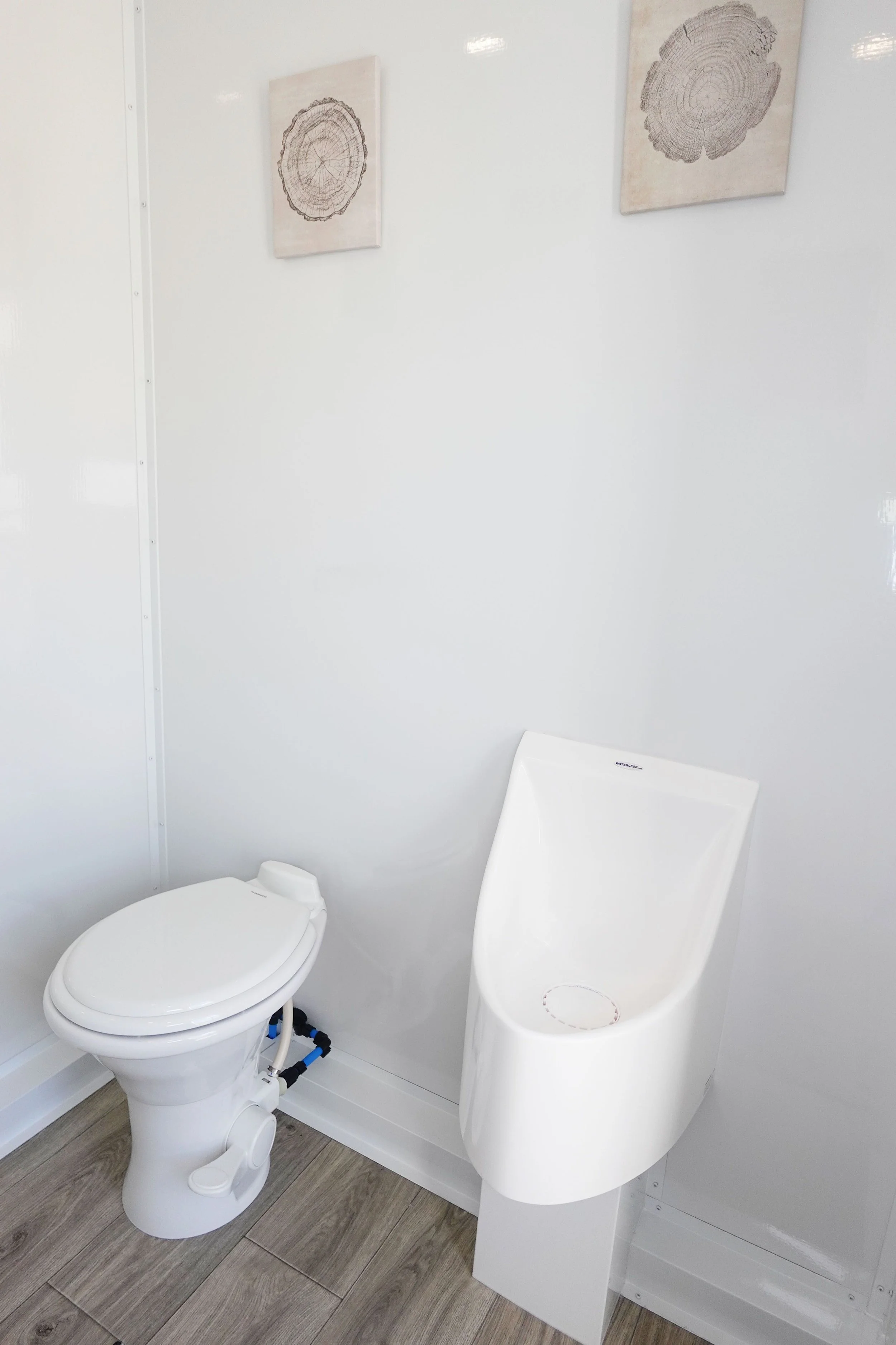 A white bathroom with a modern toilet and an electric urinal, white wall, wood-look floor, and framed wall art with tree cross-section images.
