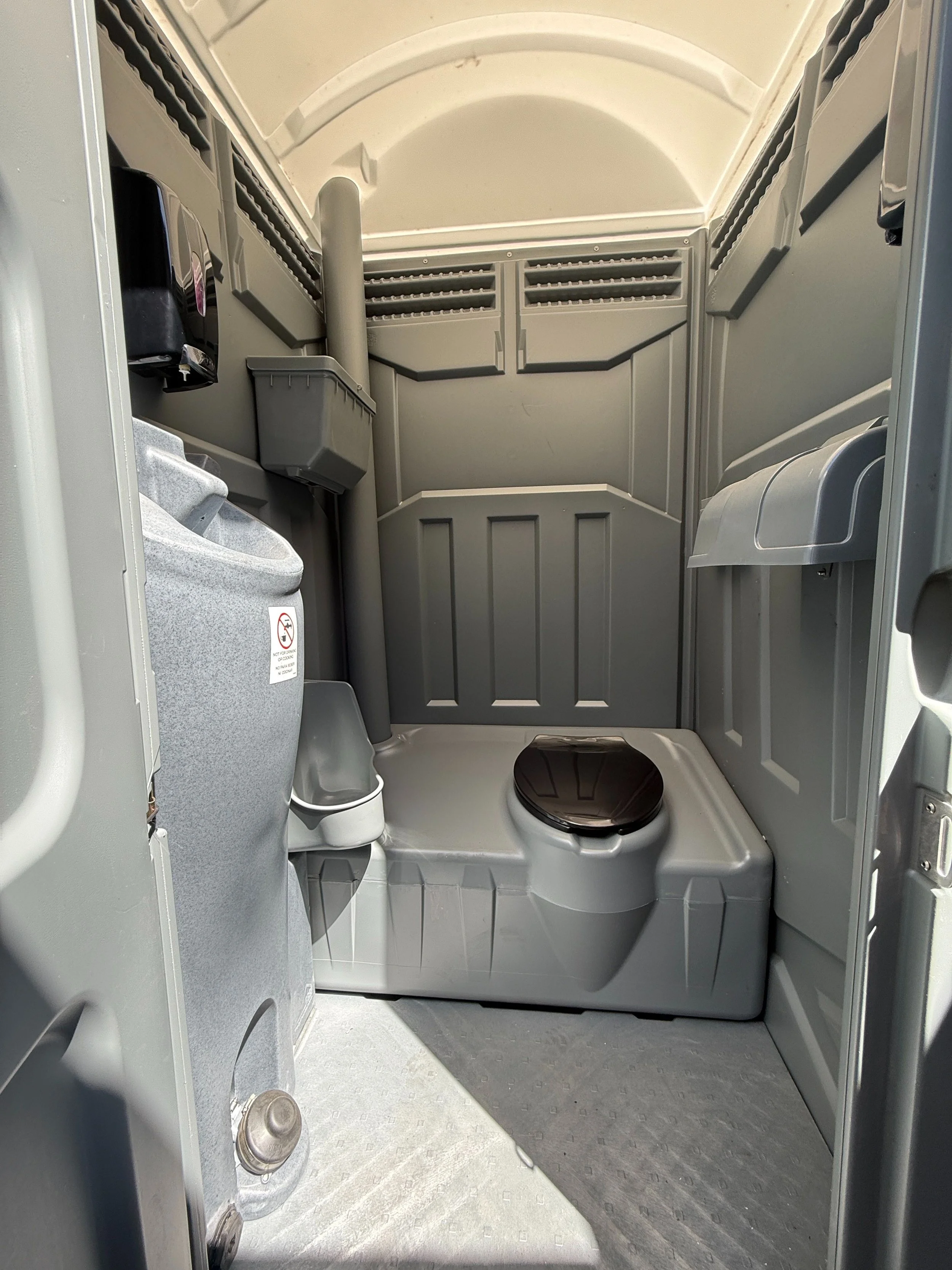 Inside of a portable restroom with gray walls, a black toilet with a closed lid, a small trash bin, and various vents and fixtures.