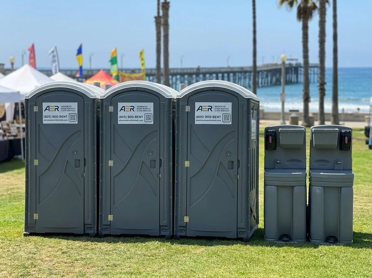 Three portable toilets and two trash bins on a grassy area near a beach with a pier, palm trees, and blue sky in the background.