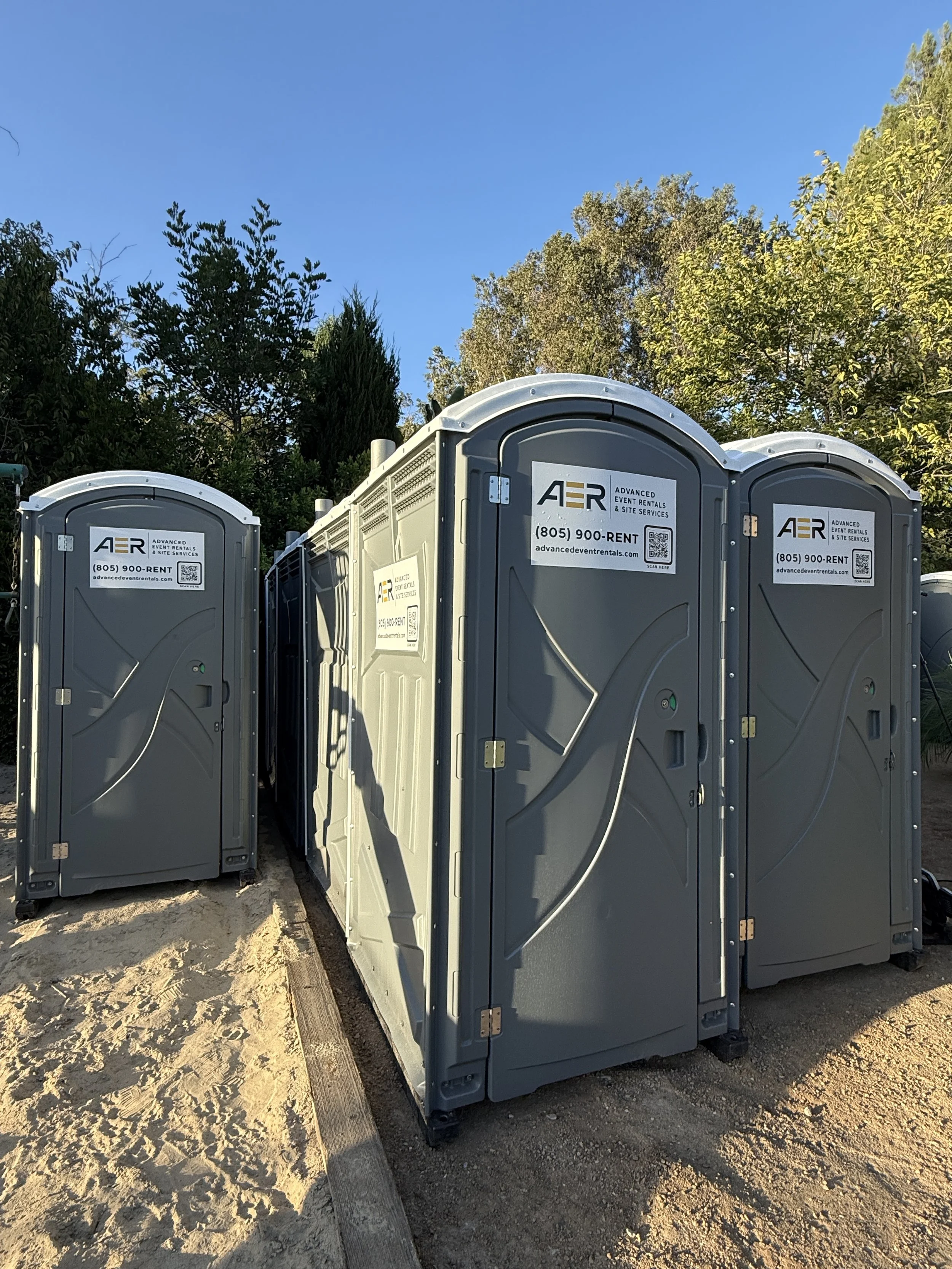 Three portable toilets with gray exteriors and white roofs, set outdoors on a dirt surface, with green trees and a clear blue sky in the background.