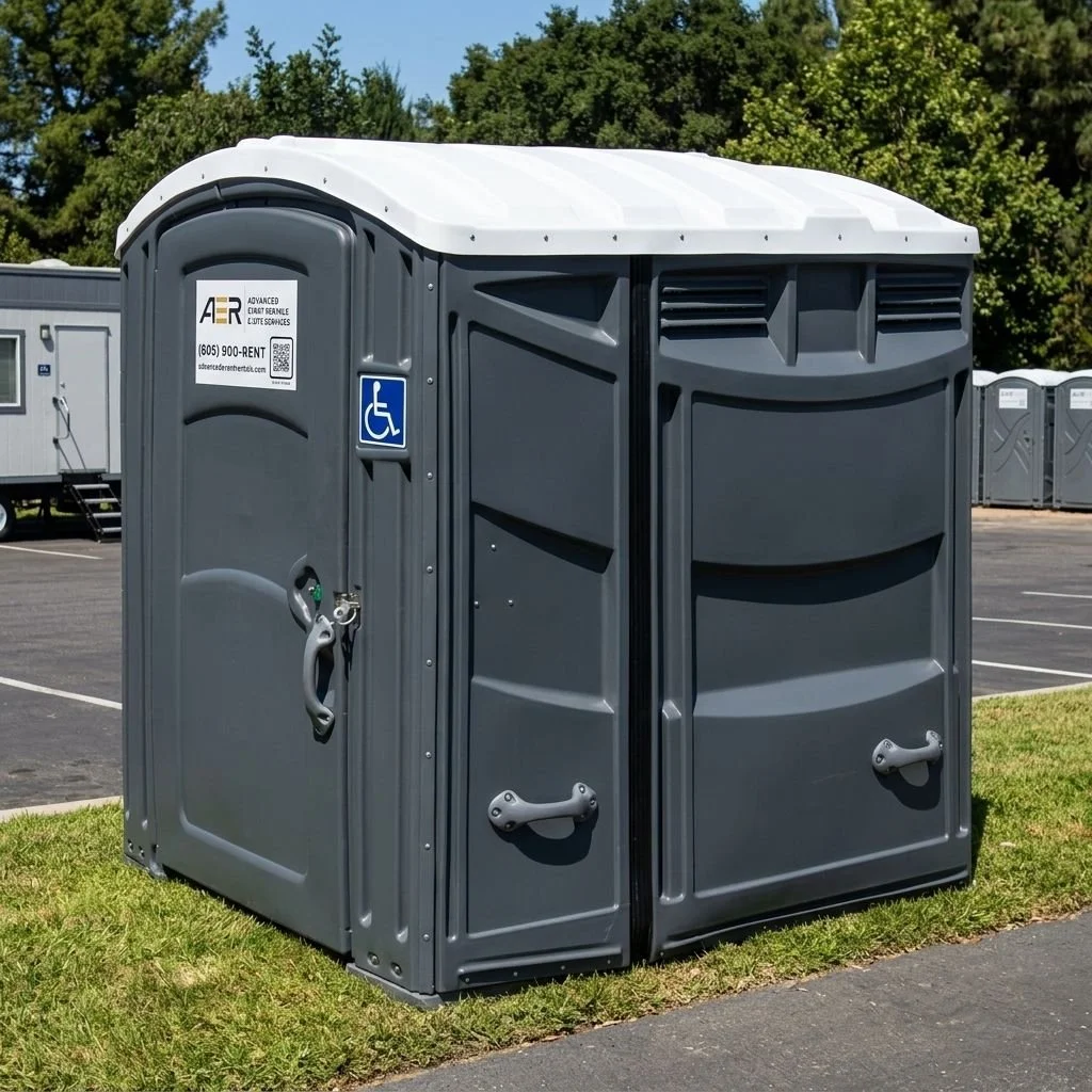 A portable, gray, wheelchair-accessible outdoor restroom with a white roof, situated on a grassy area near a parking lot with trees in the background.