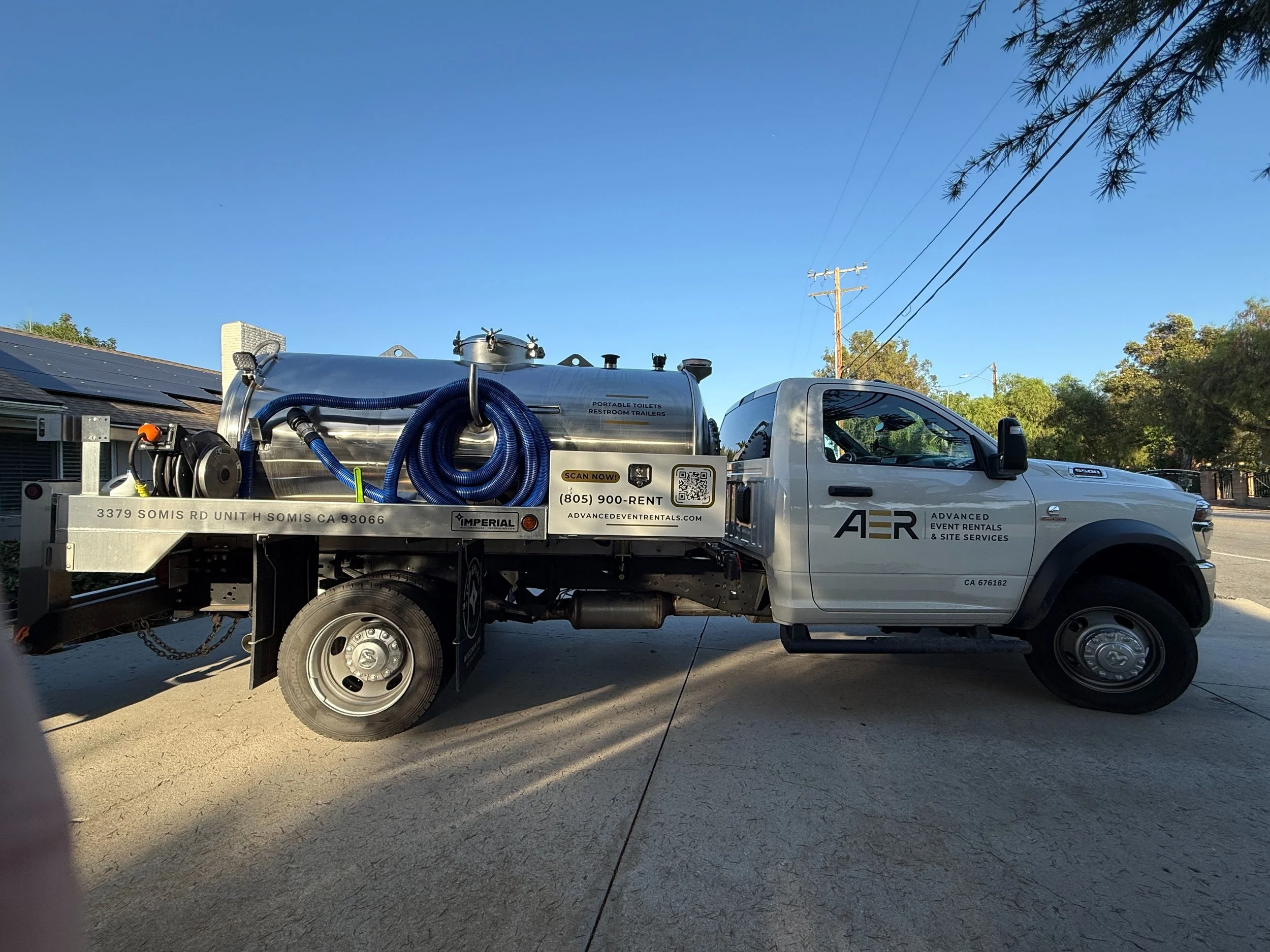 A white truck with a metal tank labeled "Portable Toilets Restroom Trailers" parked on a dirt surface. The truck has a QR code, phone number, and address on the side. The background shows a clear blue sky, trees, and utility poles.