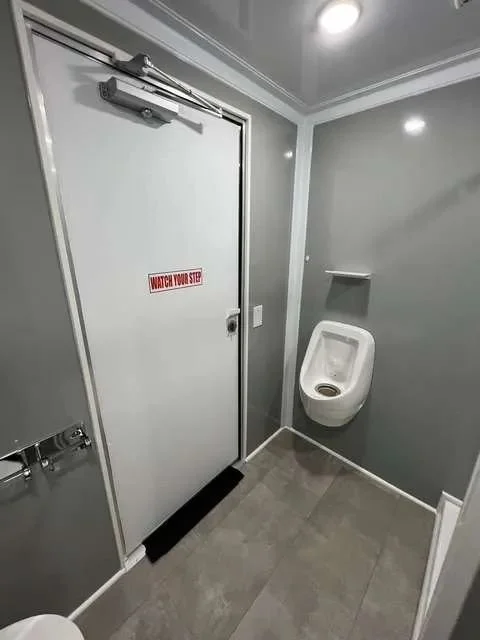 A men's restroom with a urinal on the right wall, a door with a "Watch Your Step" sign and a door closer at the top, and a small shelf above the urinal.
