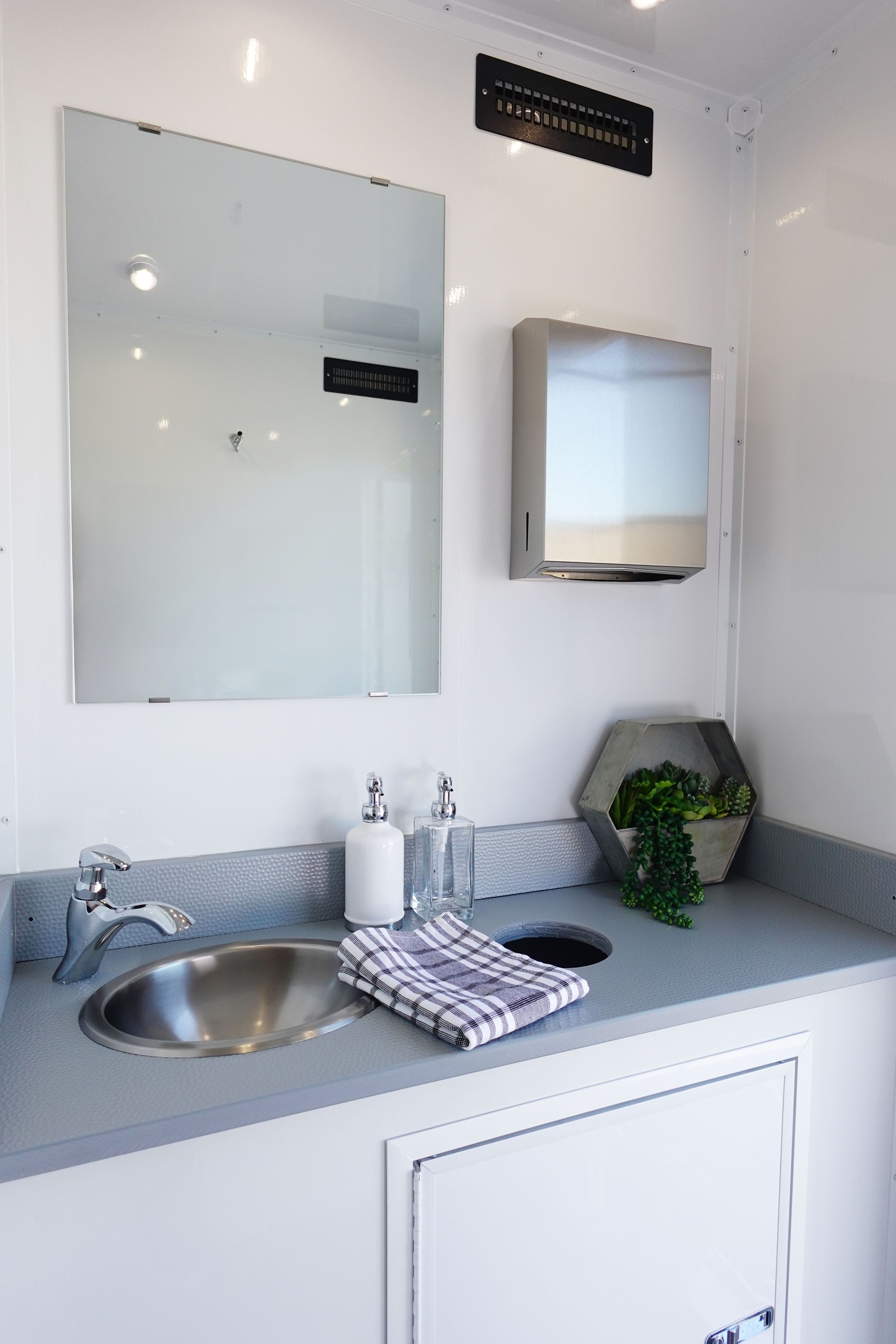 A bathroom sink with a stainless steel basin, silver faucet, soap dispenser, and a glass of hand soap on a gray countertop. A striped towel is draped over the sink. Behind the sink, there is a decorative hexagonal planter with green succulents and a rectangular stainless steel paper towel dispenser. A white wall with a mirror and black ventilation vent is visible.