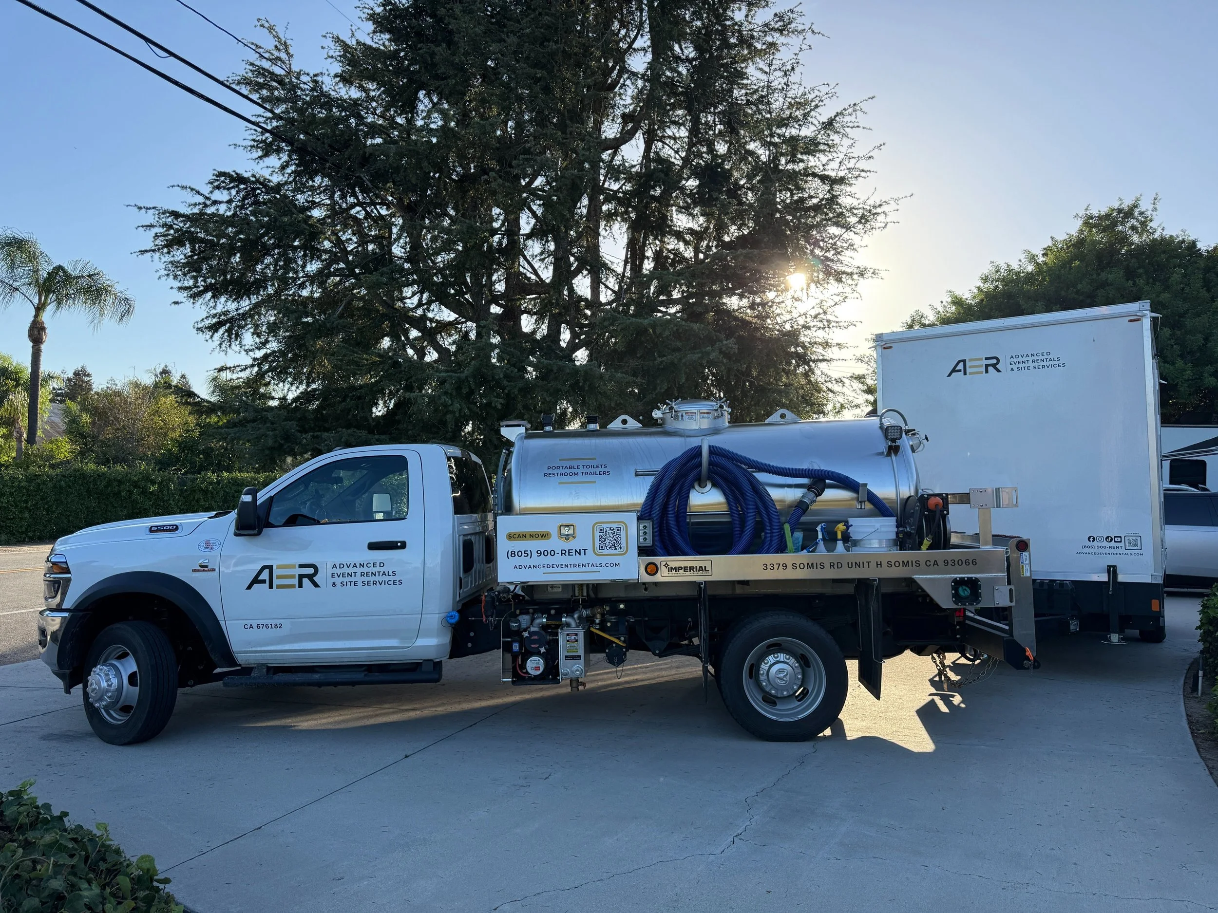 A white utility truck with equipment and hoses attached, parked in a lot with large trees and a clear sky in the background.