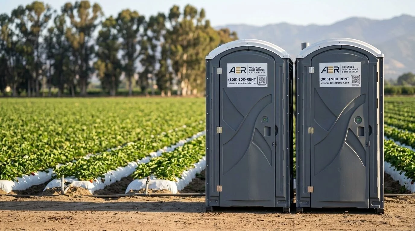 Two portable toilets on a farm field with rows of crops and trees in the background.