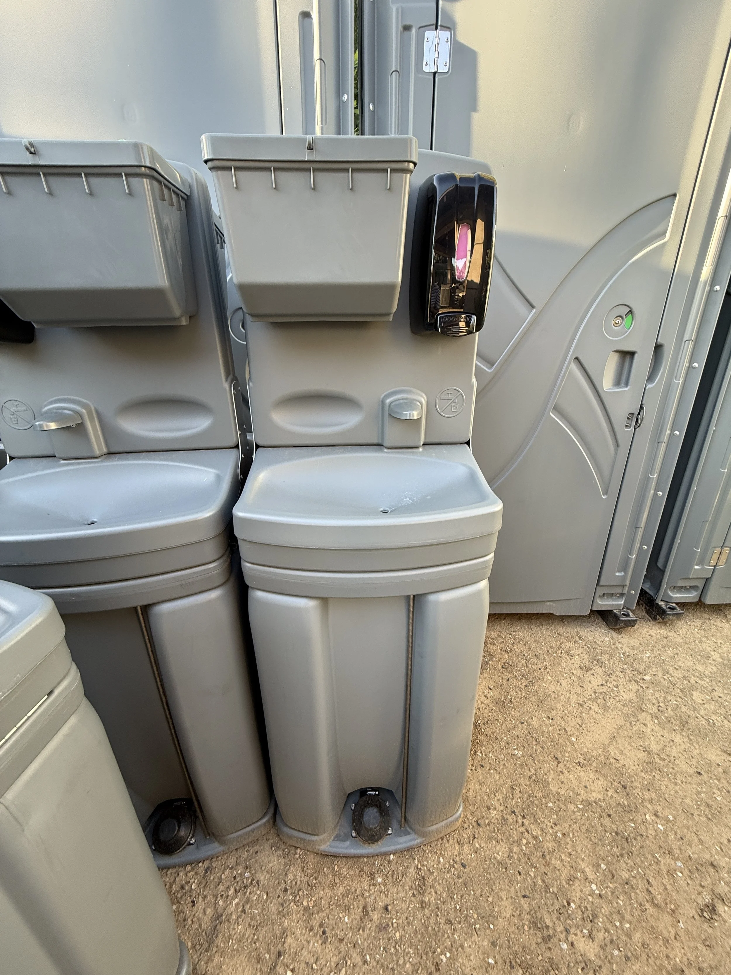 A row of gray electrical equipment or utility boxes placed outdoors on dirt ground, with some green vegetation visible behind a narrow opening.