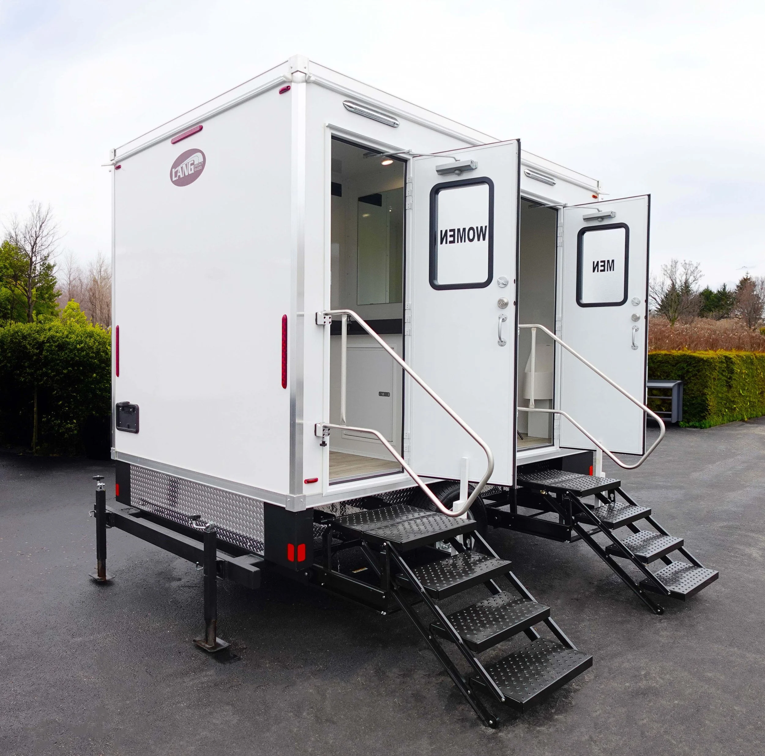 A white mobile trailer with stairs leading to the entrance, labeled "WOMEN" on the door, and featuring a logo that reads "LANG Portable" on the side. The trailer is parked on a paved surface with trees and a cloudy sky in the background.