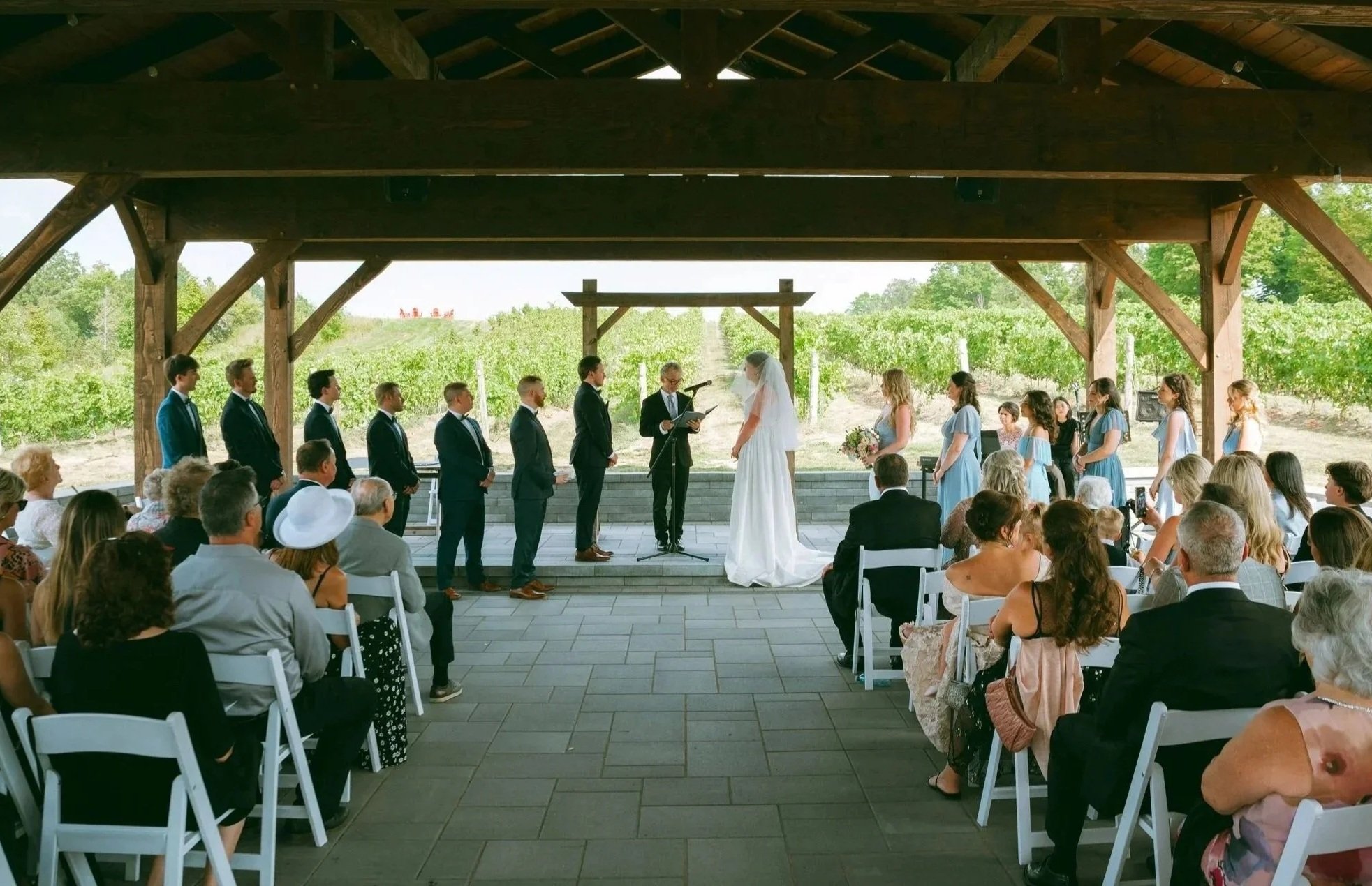 A wedding ceremony taking place outdoors under a wooden pavilion with guests seated in white chairs, the bride and groom standing in front of an officiant, with wedding party members and a scenic vineyard in the background.