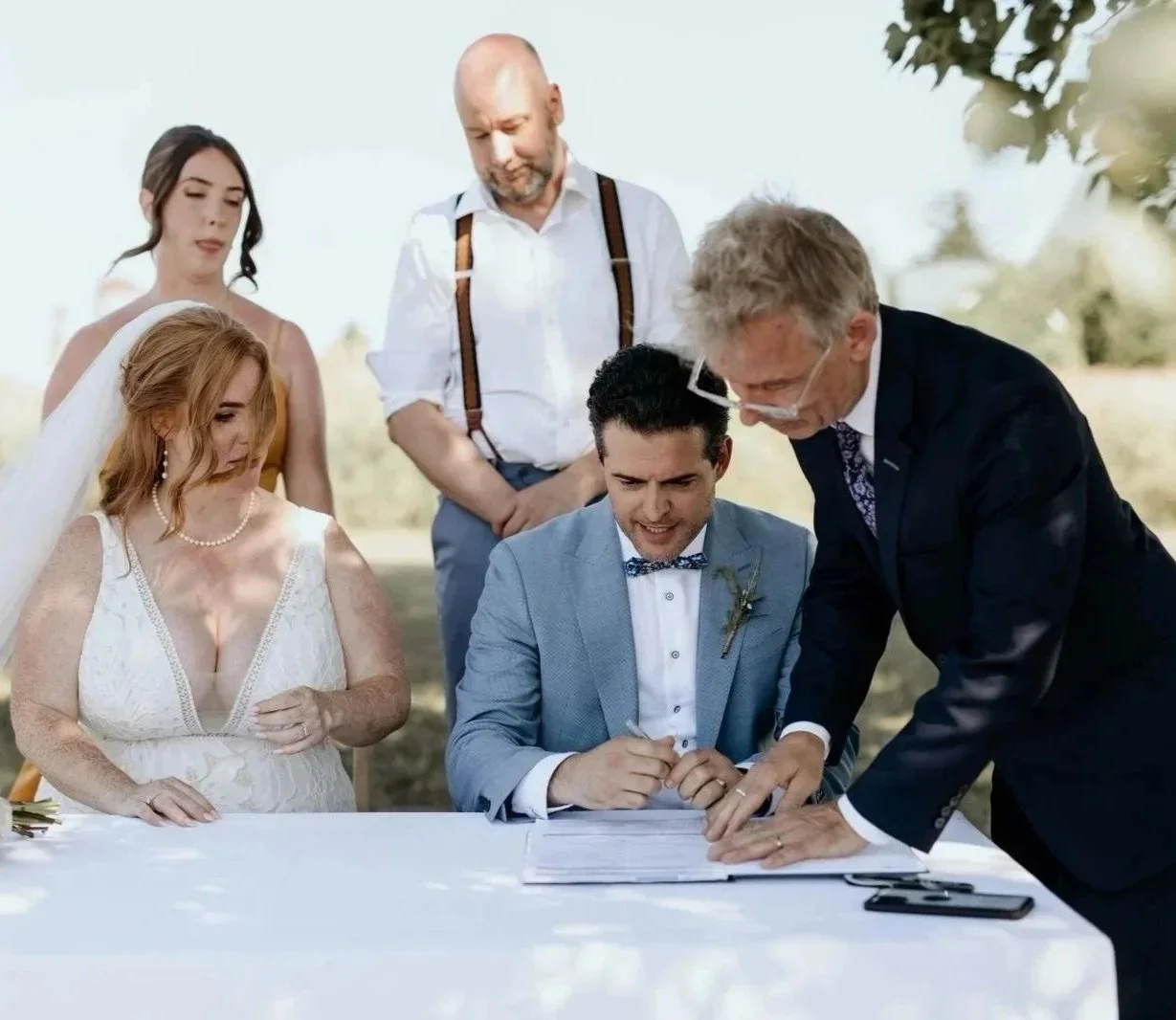 A wedding ceremony outdoors with a bride, groom, officiant, and guests signing a marriage document.