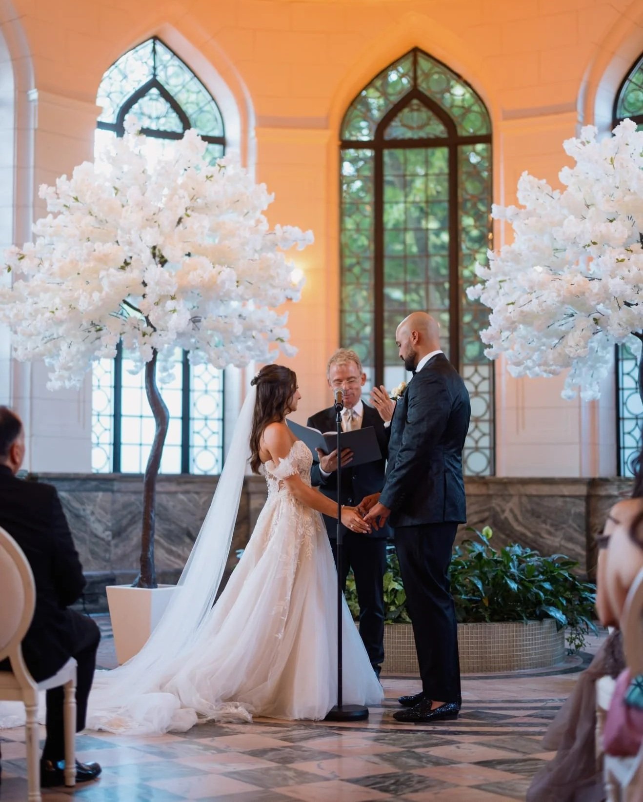 A wedding ceremony taking place indoors with a bride and groom holding hands and facing each other. An officiant, standing between them, reads from a book. The venue has large arched windows and is decorated with white flowering trees.