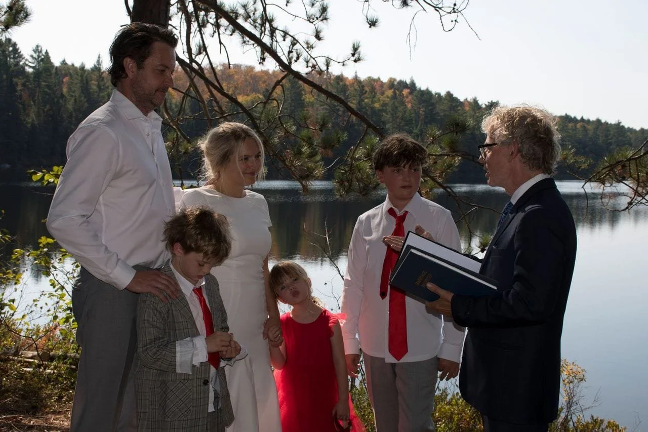 A family of four, including two children, stands by a lake near trees, listening to an older man in a suit who is holding a book and speaking to them, outdoors during daytime.