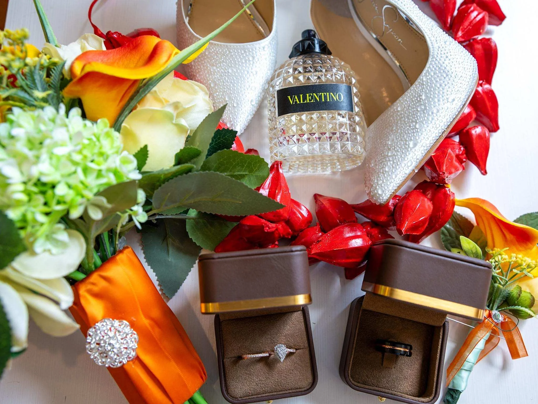 White high-heeled shoes with textured surface, a bottle of Valentino perfume, and a colorful floral bouquet with orange, yellow, white, and green flowers, arranged on a table.