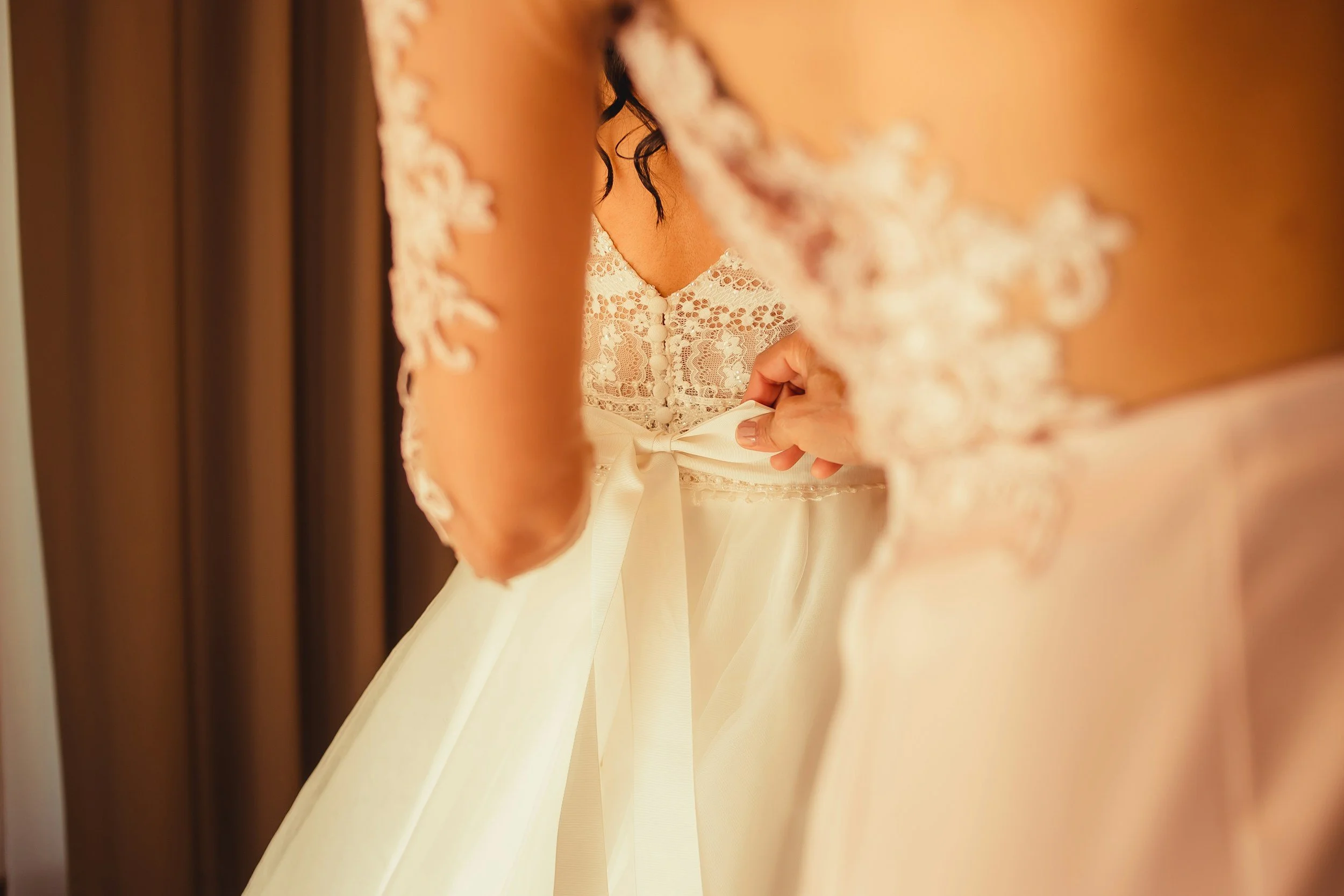 A bride fastening her wedding dress, shown through a mirror with lace details on the dress and dark, curly hair.