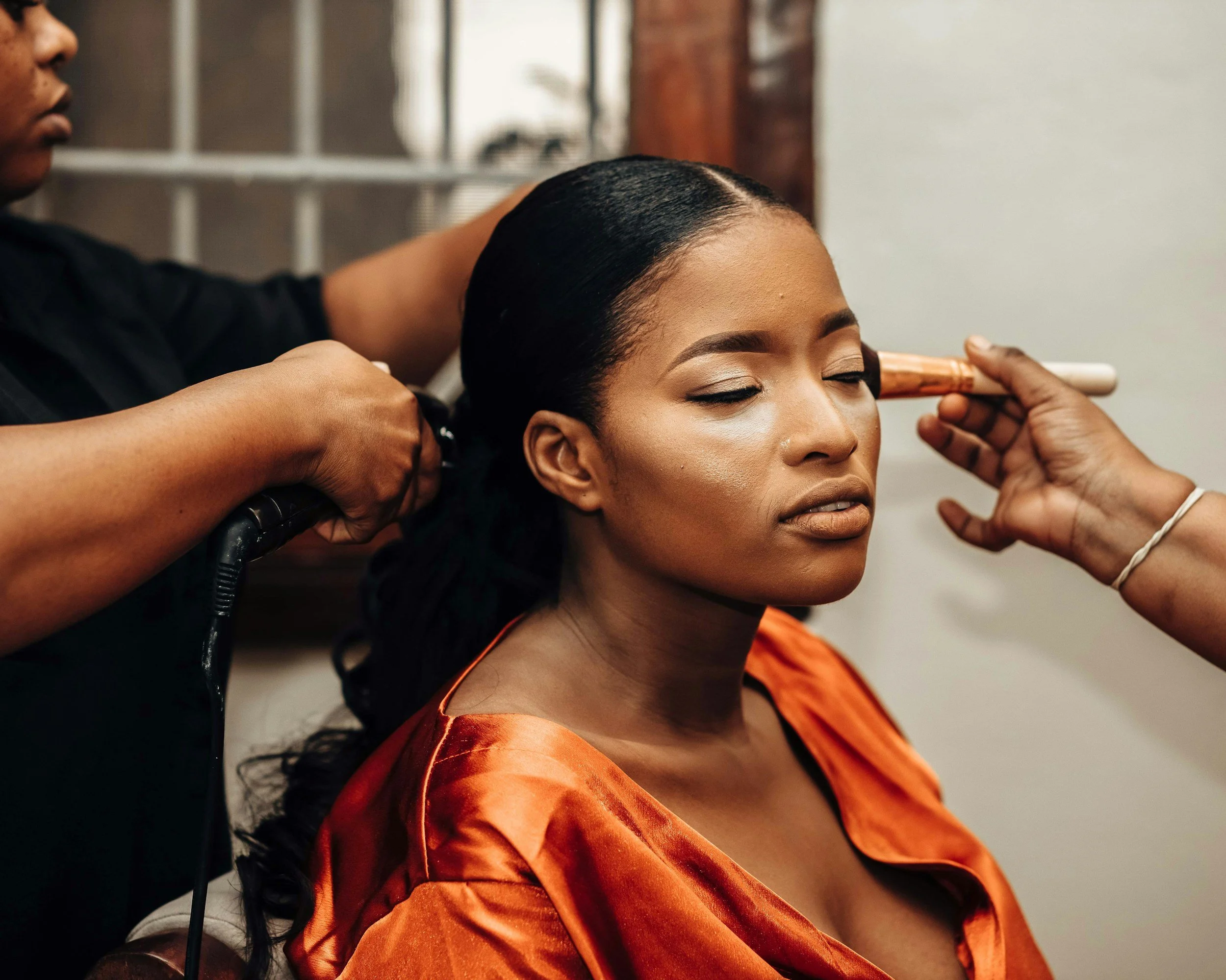 A woman with closed eyes, wearing an orange silk outfit, has her head tilted back slightly as someone does her makeup and styles her hair, which is sleek and black. The hairstylist's hands are holding a white curling iron close to her hair.