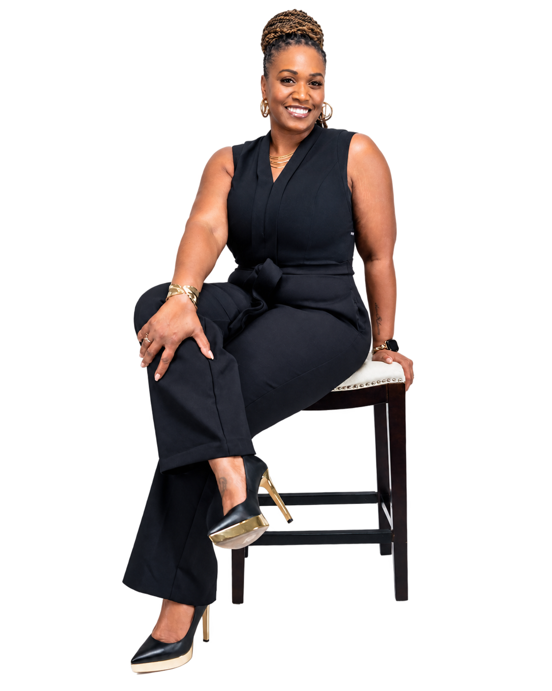 A woman sitting on a chair, wearing a black sleeveless jumpsuit, gold high heels, earrings, and jewelry, smiling at the camera.