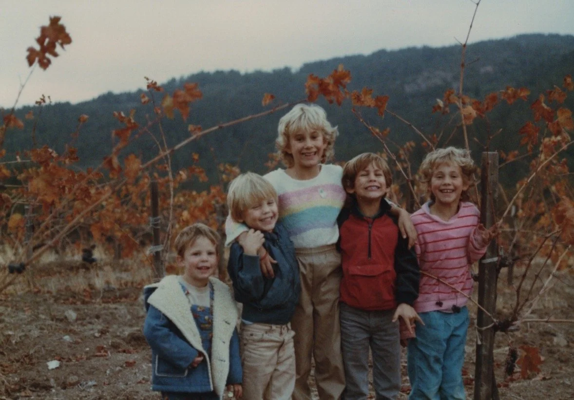 A group of five children standing outdoors among autumn foliage, smiling at the camera with mountains in the background.