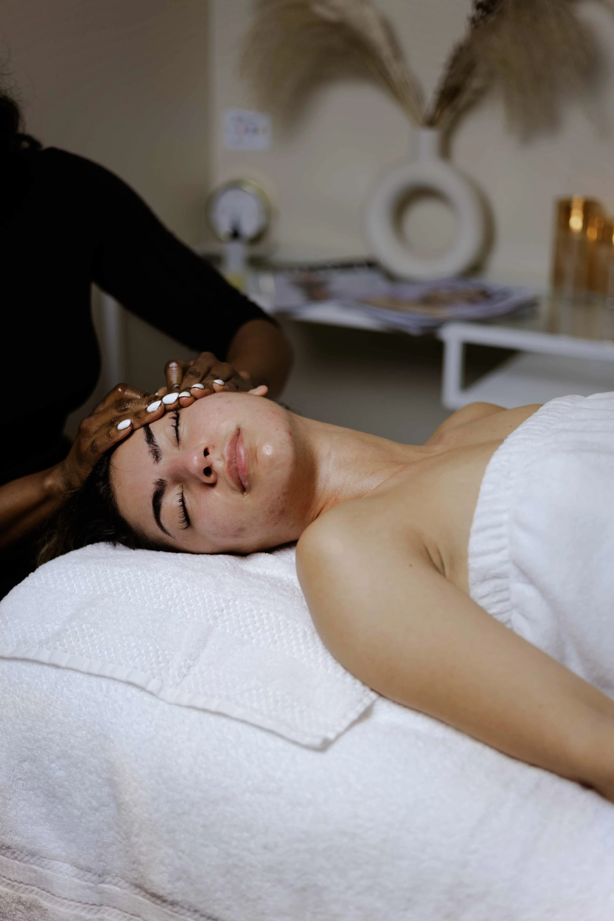 A woman lying on a massage table with her eyes closed, receiving a facial massage at Keita Beauty. Hawa Swarays hands are on her forehead, as she receives a holistic facial in Halifax Nova Scotia Canada