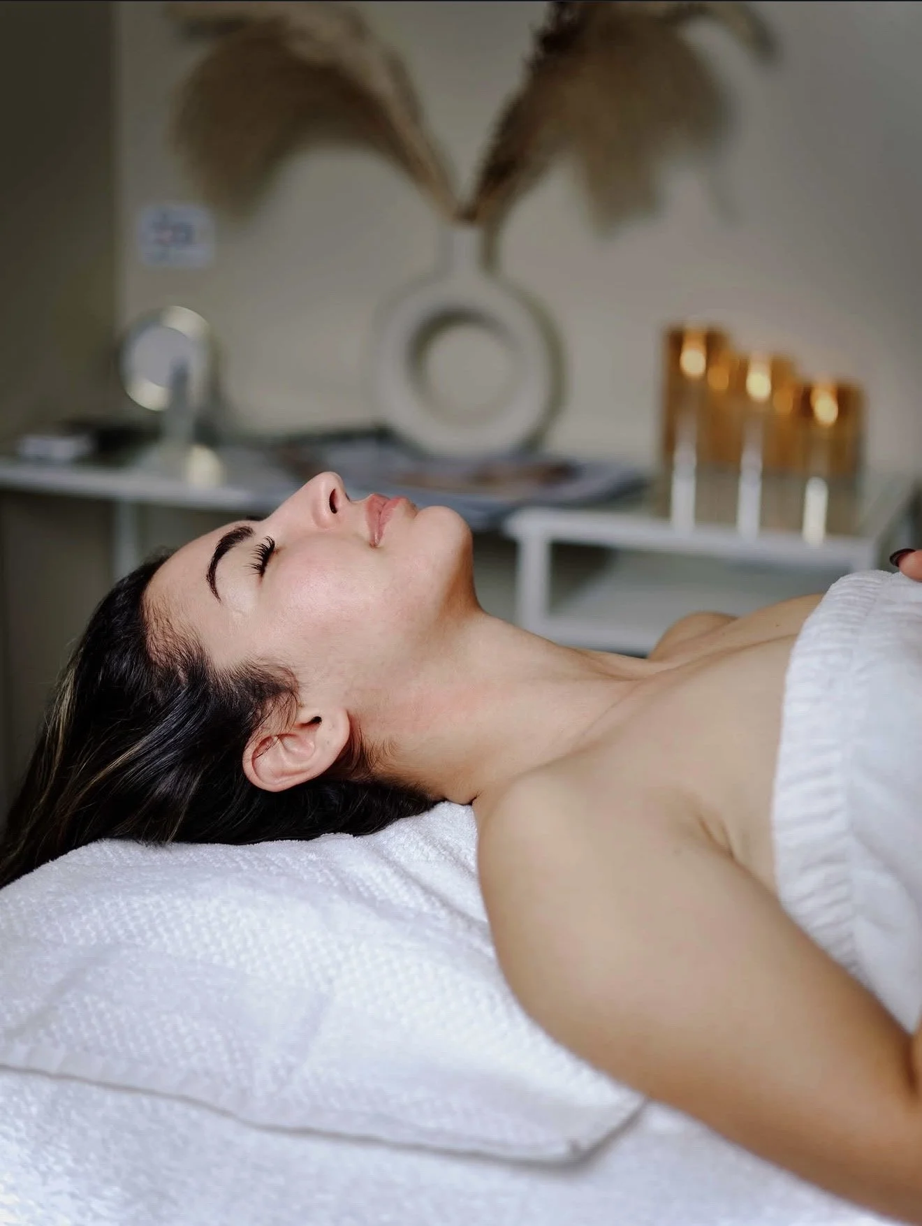 A woman lying on her back with her eyes closed in a spa or massage room at Keita Beauty, with a white towel wrapped around her chest. In the background, there is a table with four small candles and a decorative vase with dried leaves. Facial service