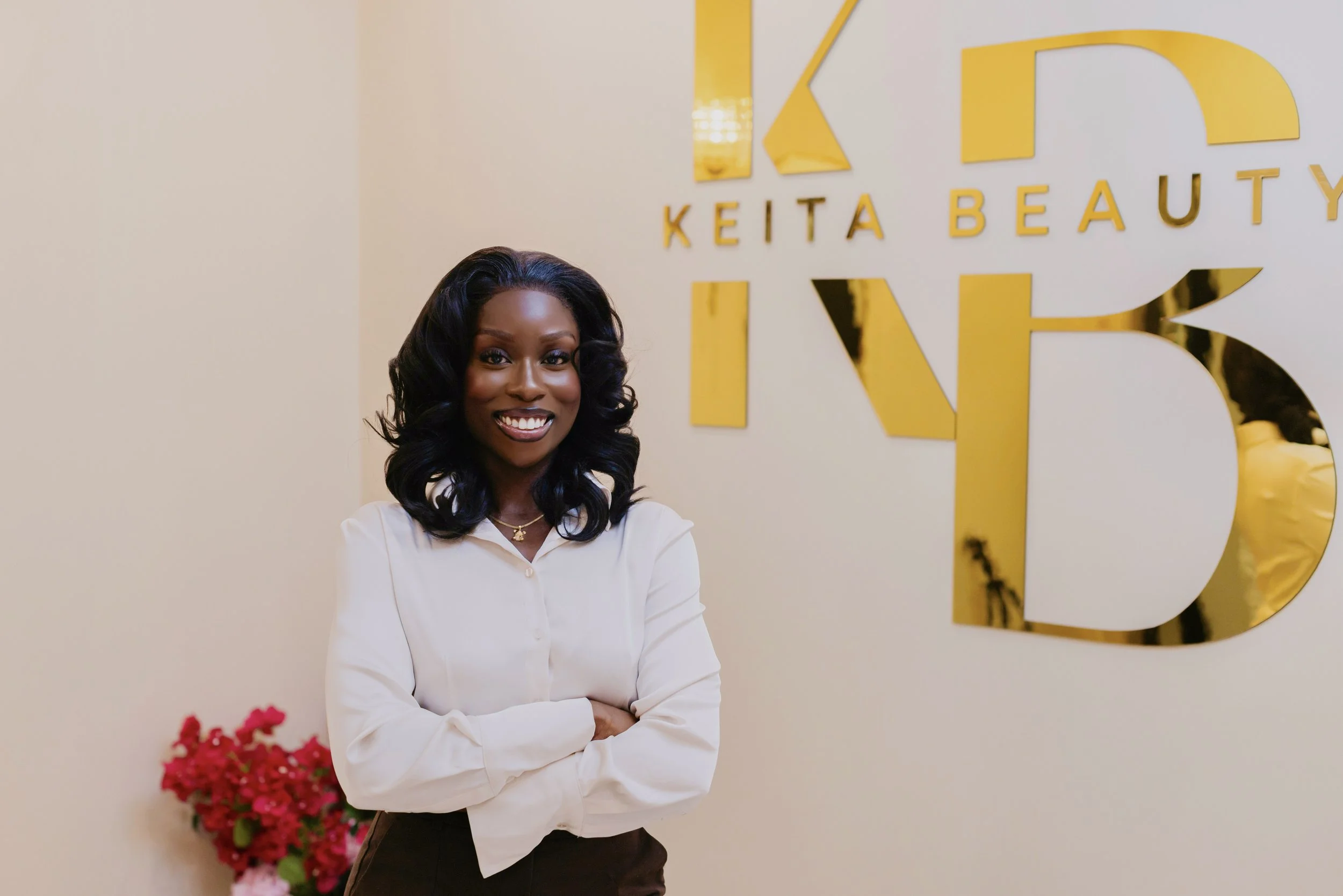 Hawa Swaray smiling with dark, wavy hair wearing a white blouse and black pants stands with her arms crossed in front of a wall with a gold sign that reads "KEITA BEAUTY" and a large stylized letter "K" in Halifax Nova Scotia Canada