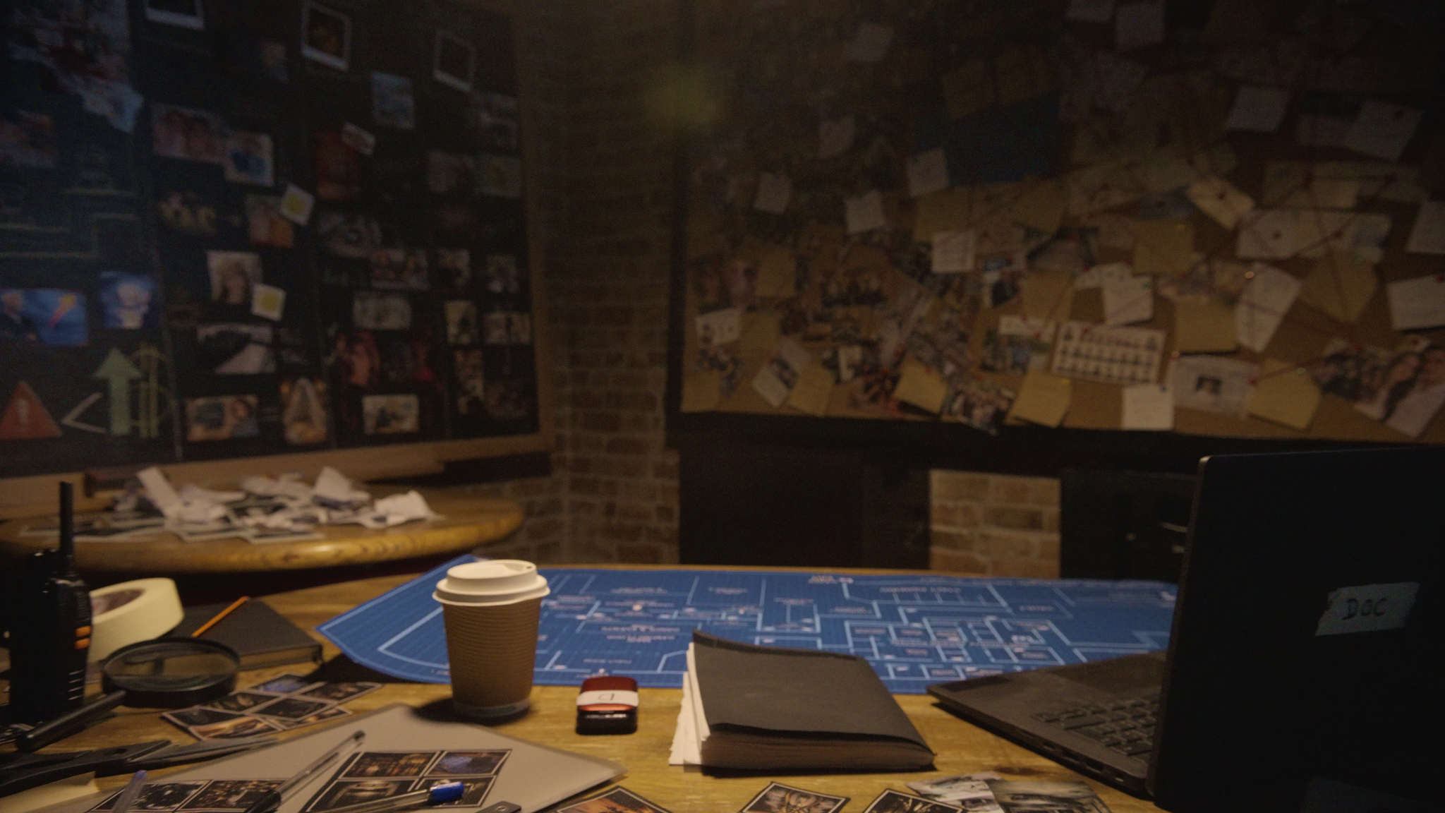 Mesa de juego con planos, fotografías, libreta y una taza de café, en un ambiente con paredes de ladrillo y cartelera con fotos y notas.