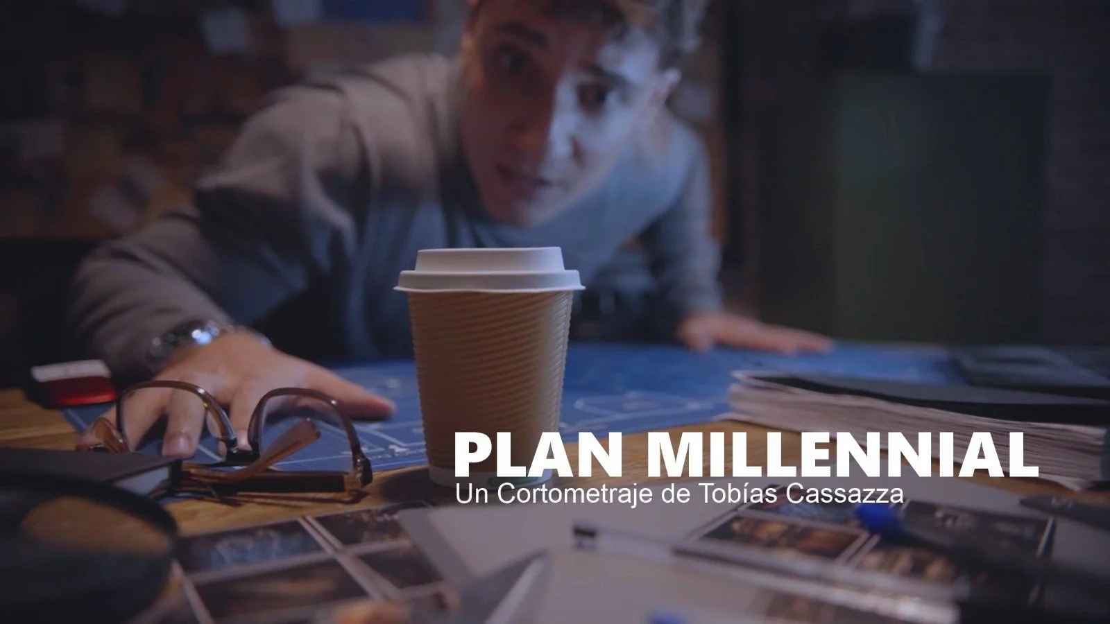 Un hombre se acerca a una mesa con documentos, una taza de café de cartón, gafas y fotografías, en un ambiente de oficina o estudio, en un cortometraje titulado 'Plan Millennial' dirigido por Tobías Cassazza.