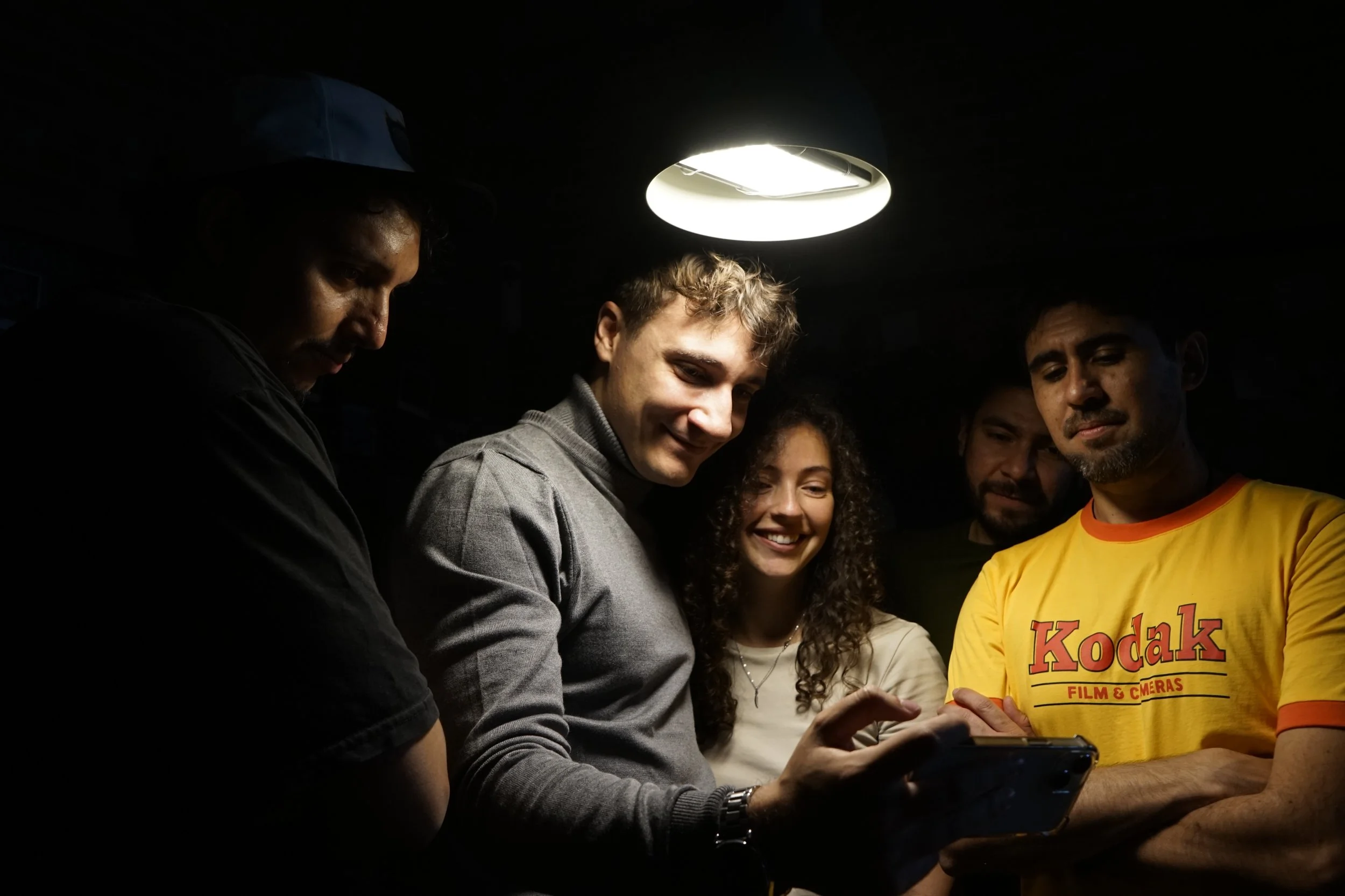 Grupo de personas observando una pantalla o teléfono móvil en un entorno oscuro. Se ven sonrisas y atención en sus rostros, con un foco de luz brillante sobre sus cabezas.