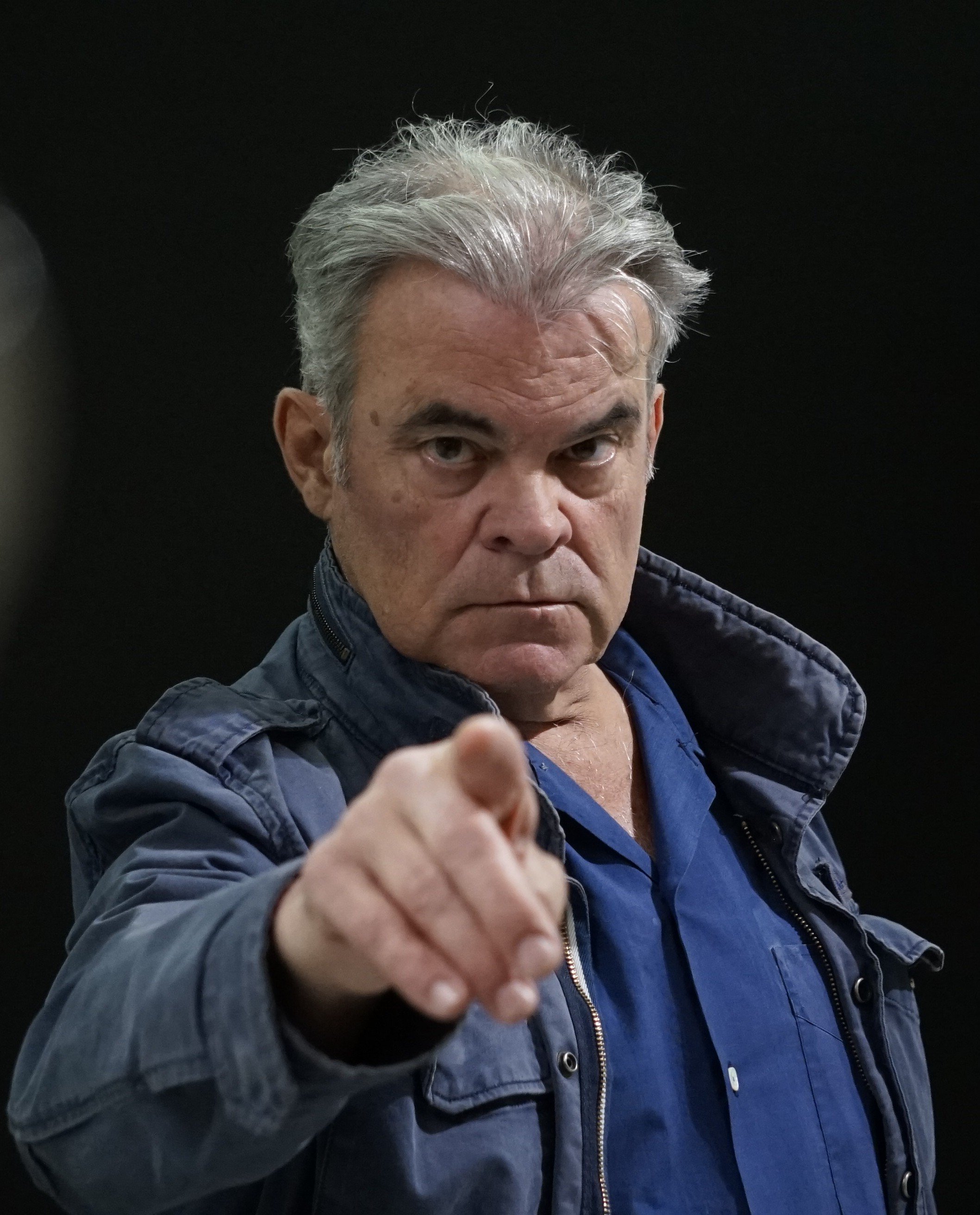 Retrato de un hombre mayor con cabello gris, vistiendo una chaqueta azul y una camisa del mismo color, señalando hacia la cámara con expresión seria en un fondo negro.