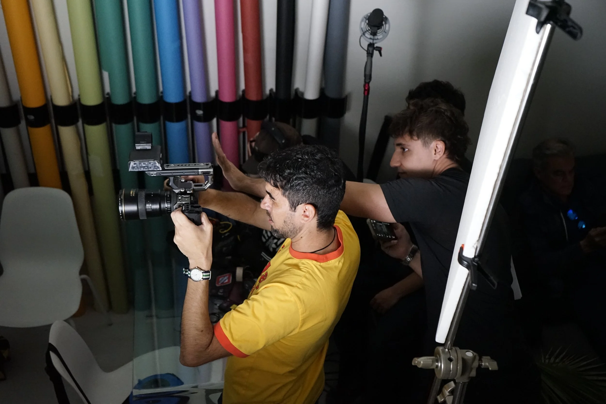 Dos hombres en un estudio fotográfico manipulan un equipo de cámara y una luz, con tubos de colores en la pared de fondo.