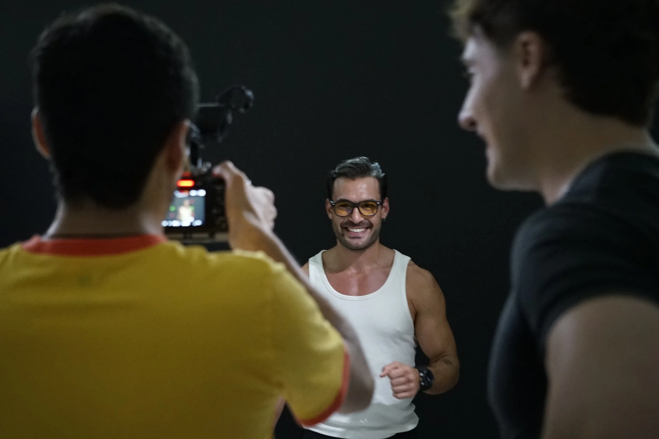 Un hombre sonriendo, usa gafas con lentes anaranjados y camiseta blanca, en el fondo un fondo oscuro, rodeado de dos personas capturando una foto o video de él.