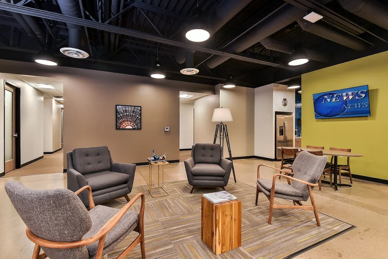 A modern waiting area with four chairs arranged around a small wooden table, a floor lamp, a television on a yellow wall displaying a news broadcast, and a hallway leading to other rooms.