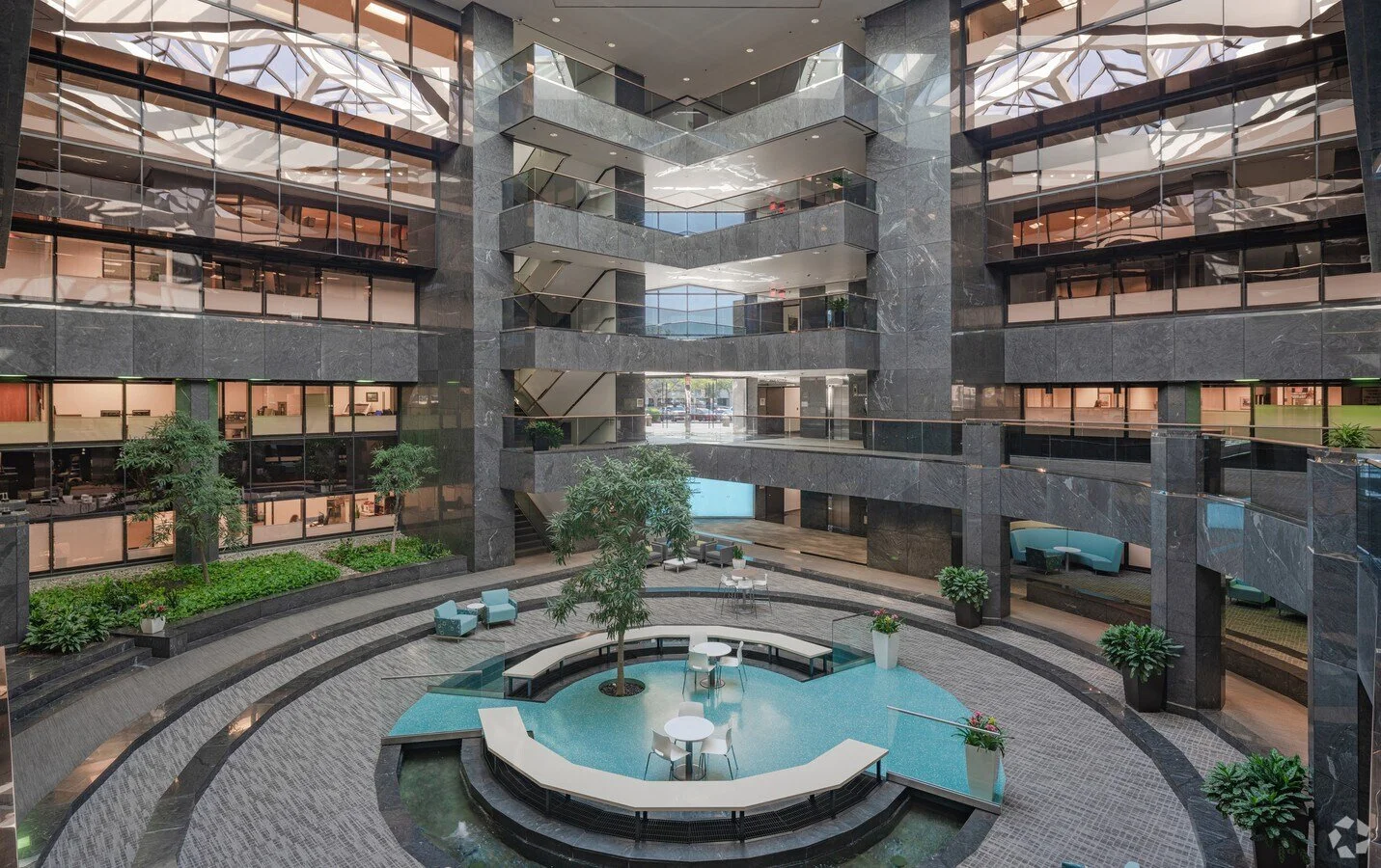 Interior view of a modern multi-story office building with an open atrium, featuring glass walls, plants, and seating areas around a central water feature.