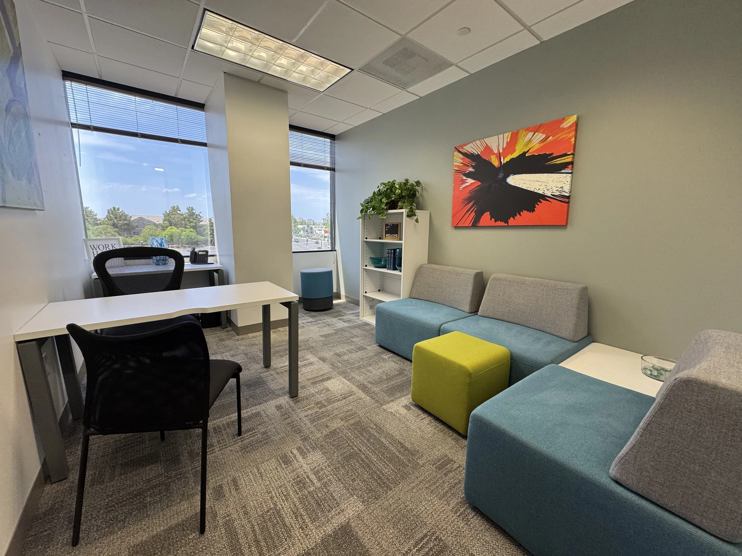 An office waiting area with a blue and gray sofa, a white bookshelf, a green ottoman, a desk with a black chair, large windows with blinds, a colorful abstract painting, potted plant, and a carpeted floor.