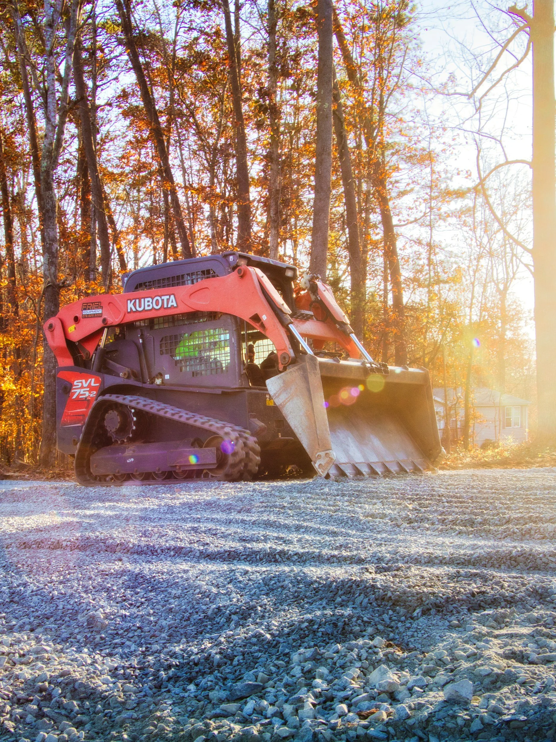 A red Kubota mini-excavator working on a gravel surface in a wooded area during sunset.