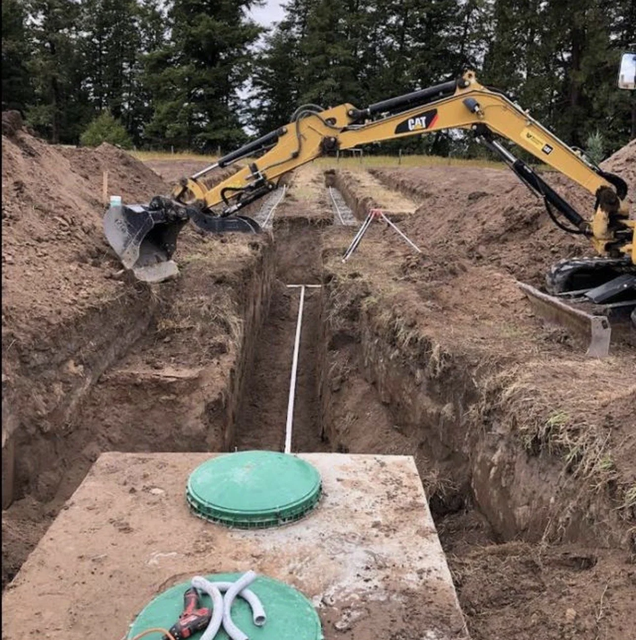 Construction site with an excavator digging a trench for underground utilities, with pipes and a large green access cover in the foreground.