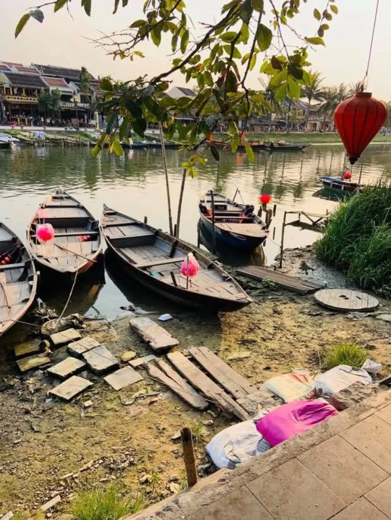 A riverside scene with several small wooden boats moored along the shore, some with pink lanterns inside. Overhanging branches with green leaves, a red lantern hanging, and a row of buildings across the river in the background. The shoreline has dirt, bricks, and sacks, with a small wooden dock extending into the water.