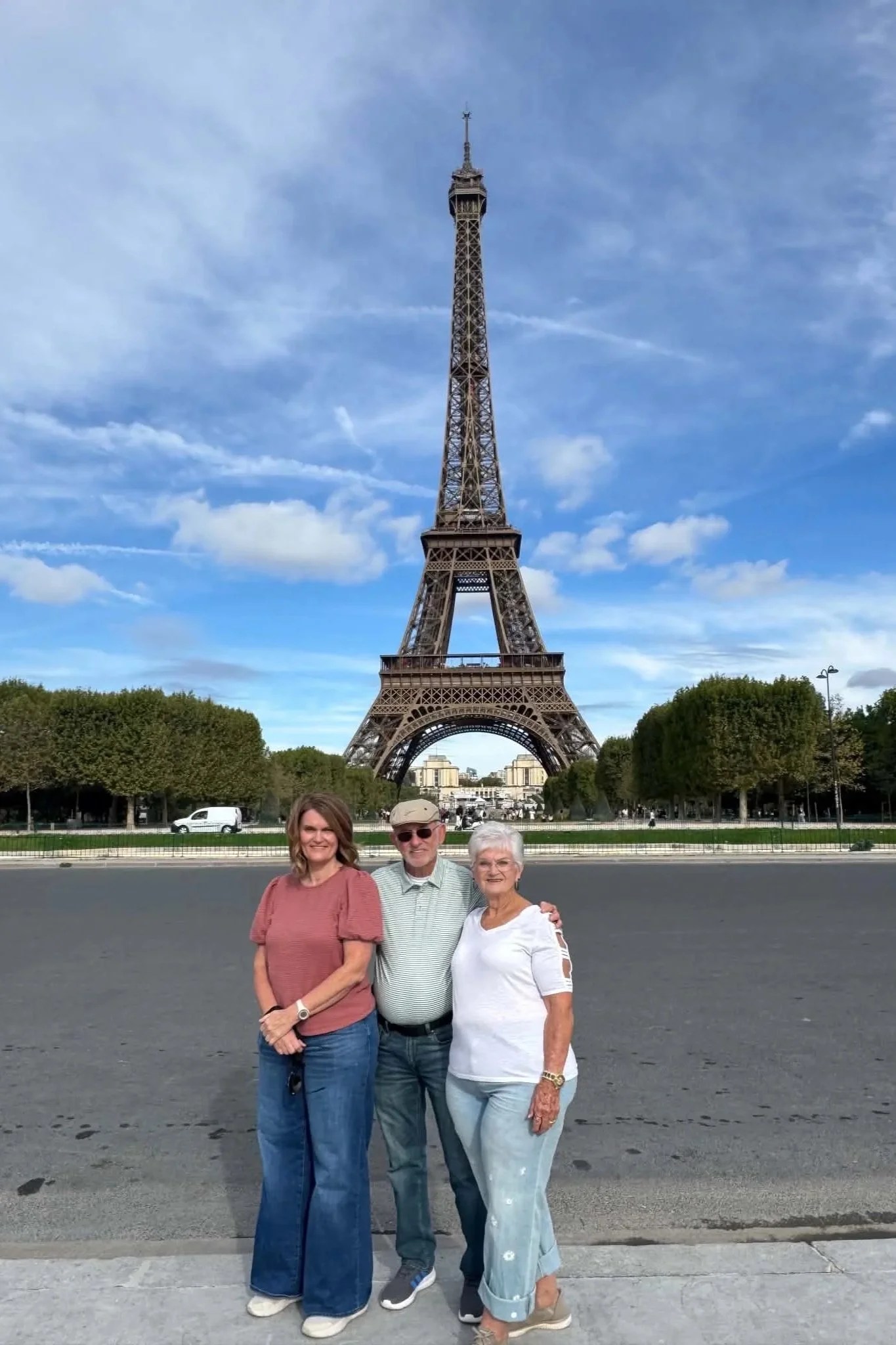Three people standing in front of the Eiffel Tower in Paris, France on a clear day. Two women and one man posing for the photo, with trees and buildings in the background.