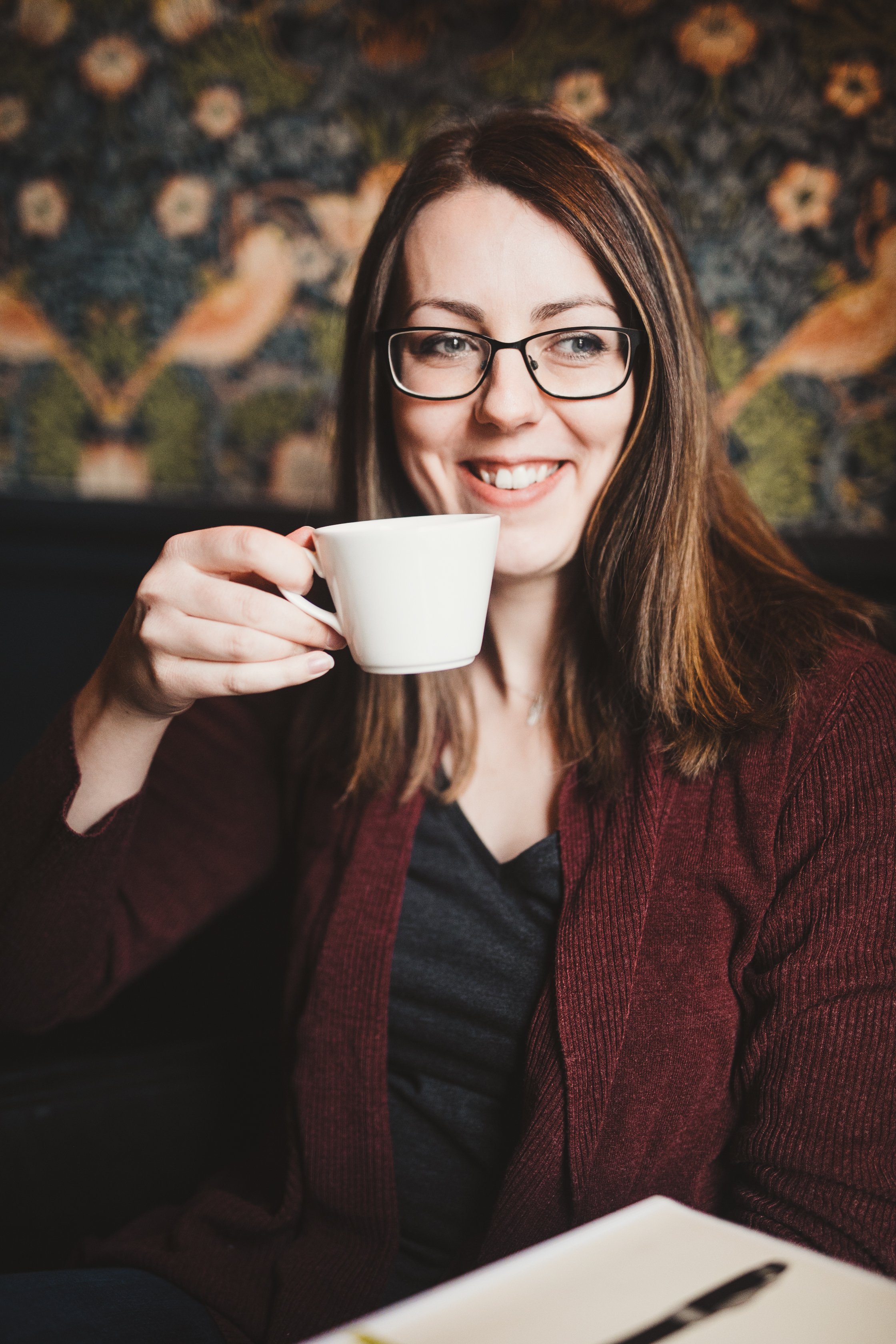 A woman with brown hair and glasses smiling and holding a white coffee cup in a cozy setting with a William Morris wallpaper background.