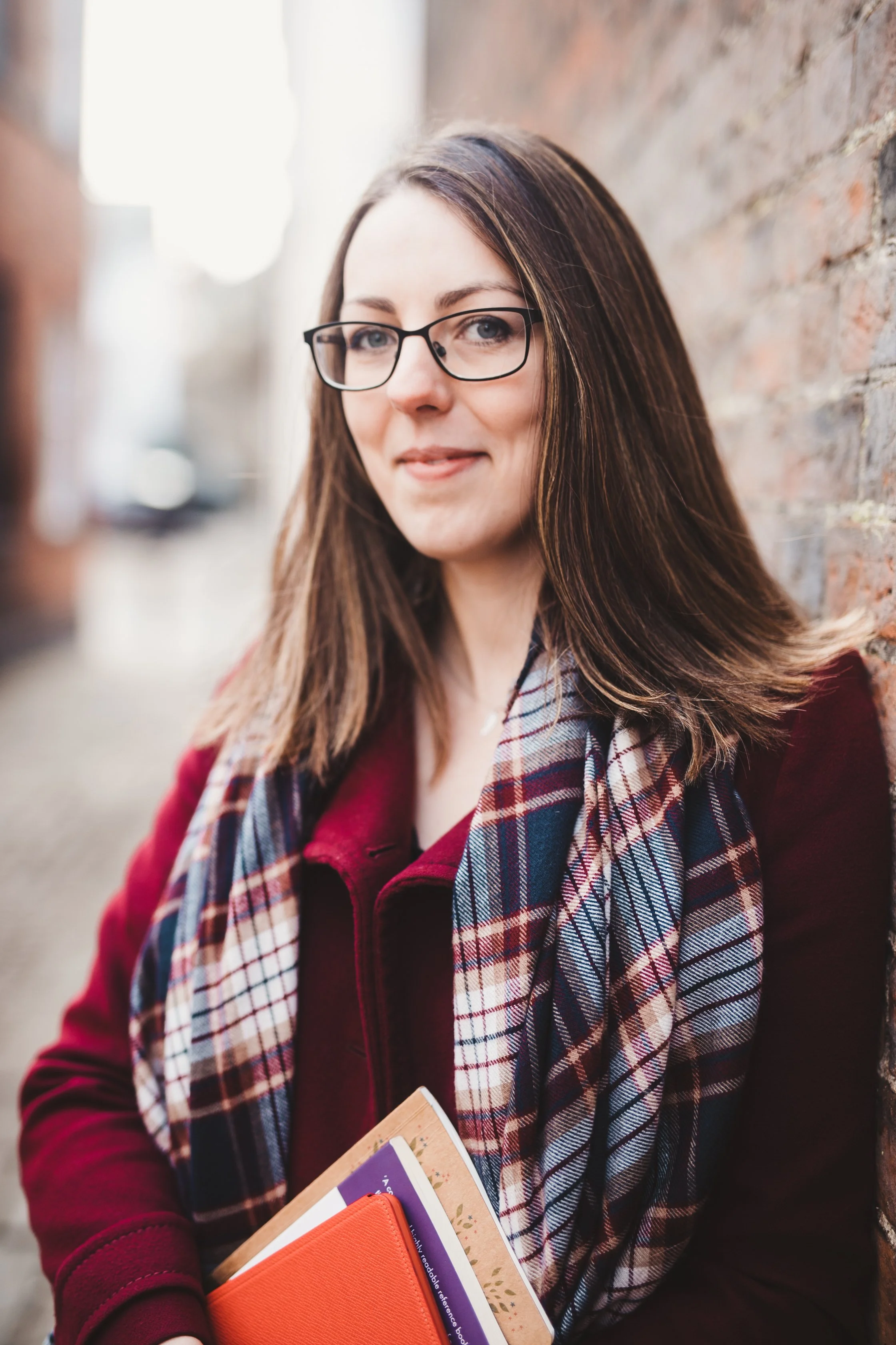 A woman with brown hair, glasses, and a plaid scarf stands against a brick wall, holding books and folders.
