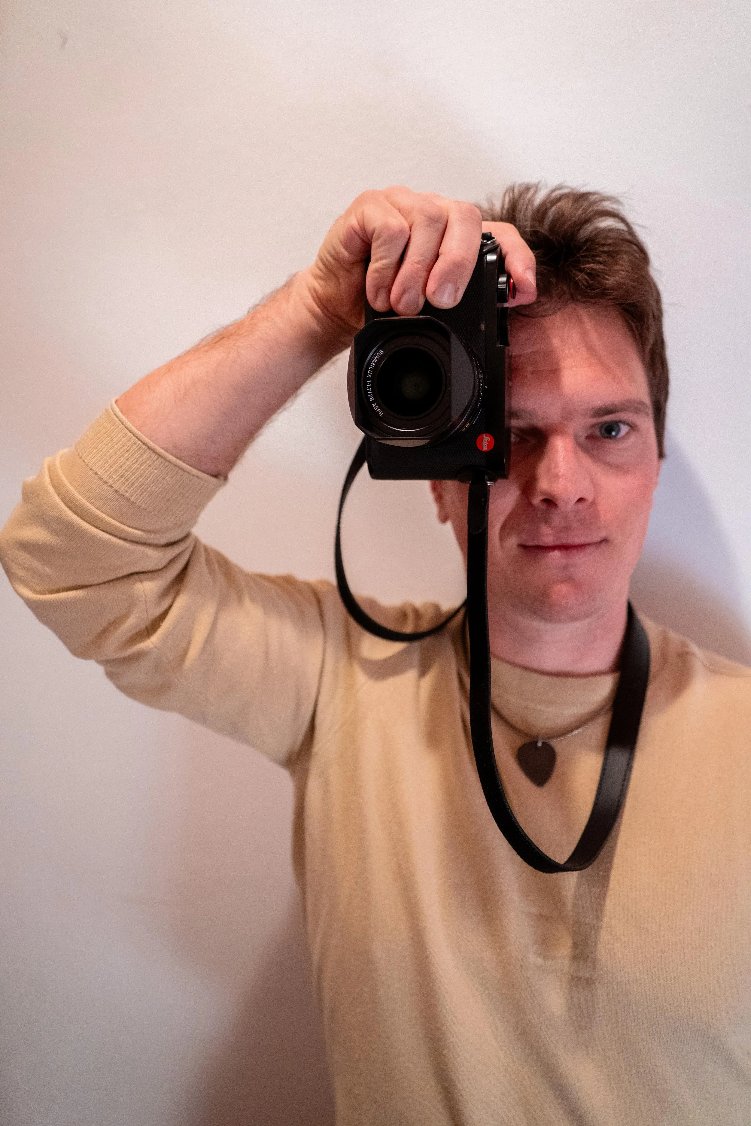 Ein junger Mann mit braunen Haaren und blauer Augen, trägt einen beigen Pullover, hält eine Kamera vor sein Gesicht und fotografiert, vor einem weißen Hintergrund.