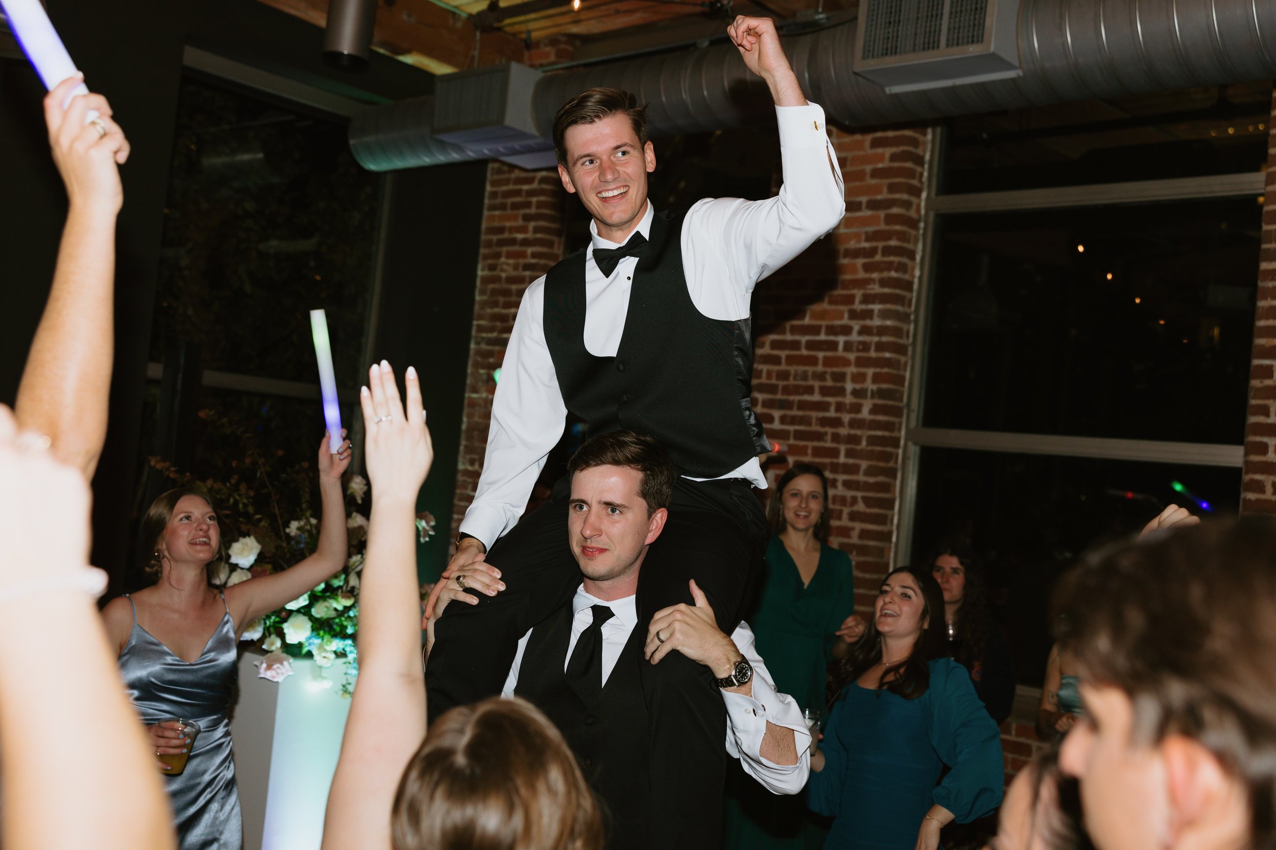 A man in a wedding tuxedo is in the air around his groomsmen at Huguenot Loft in Greenville, SC as his wedding reception enjoying music from his wedding DJs.