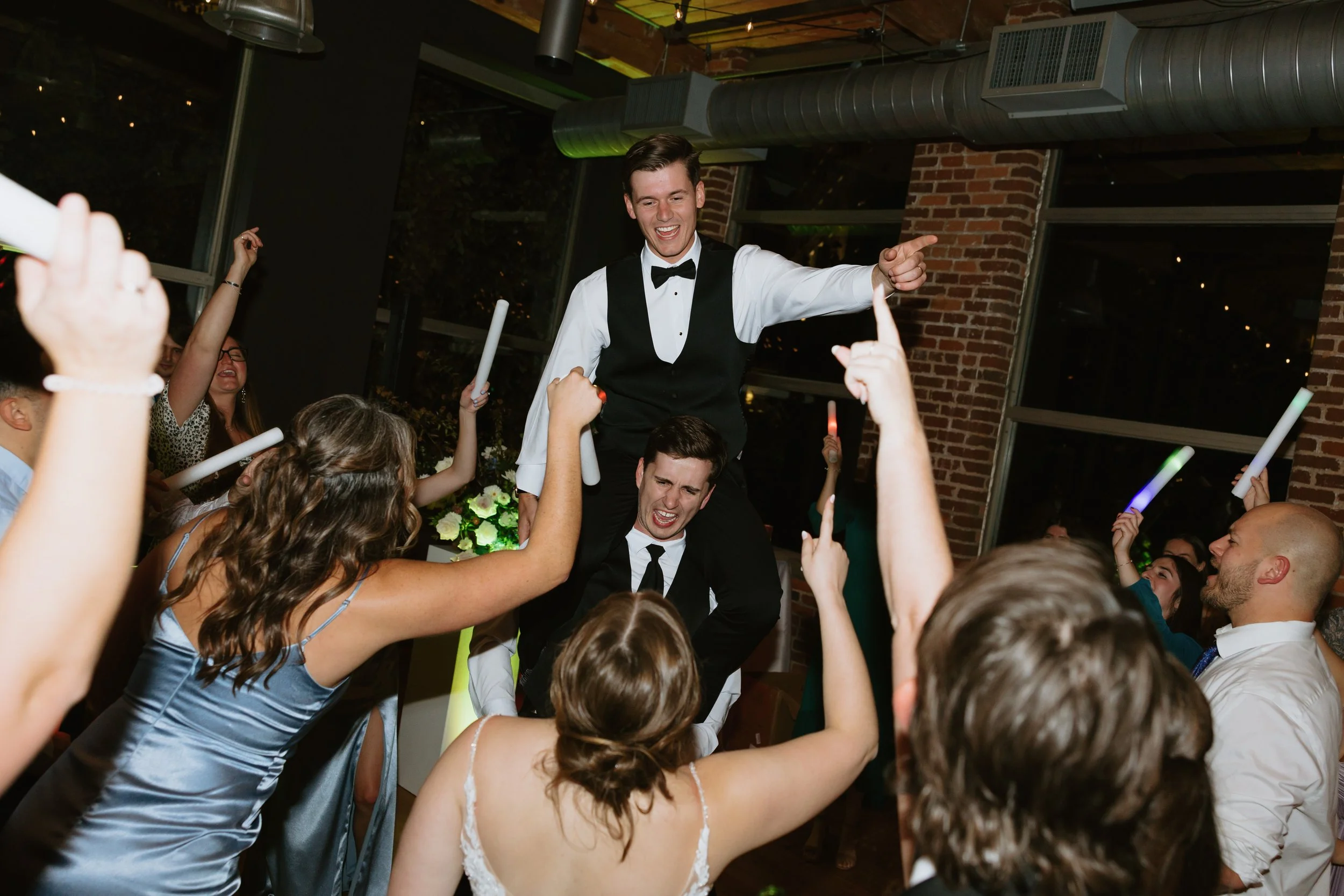 A man in a wedding tuxedo is in the air around his groomsmen at Huguenot Loft in Greenville, SC as his wedding reception enjoying music from his wedding DJ