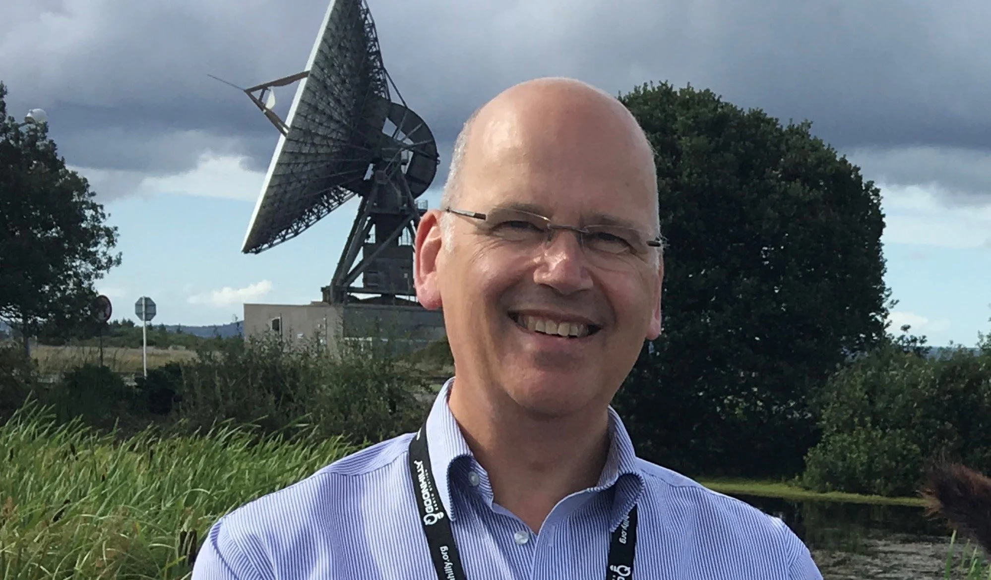 Ian Jones, OBE, Founder of Goonhilly Earth Station is a smiling man with glasses and a blue striped shirt stands outdoors in front of a large satellite dish and trees under a cloudy sky.