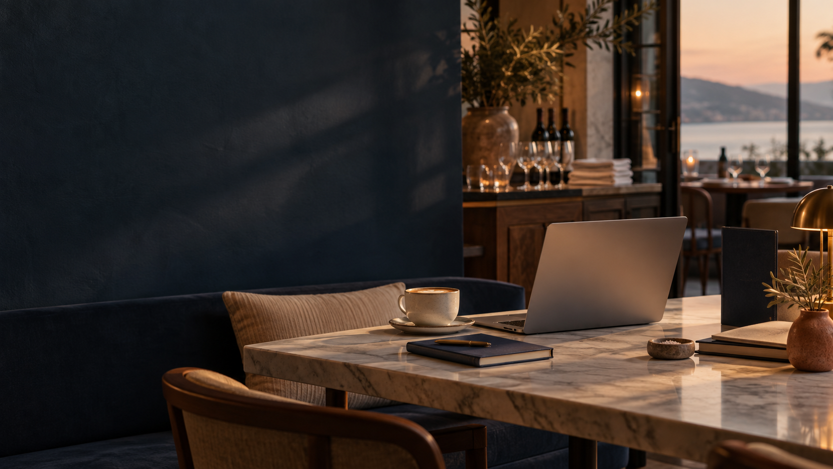 A cozy, well-lit dining space with a marble table, laptop, notebook, cup, and decorative plants, overlooking a sunset view through large windows.