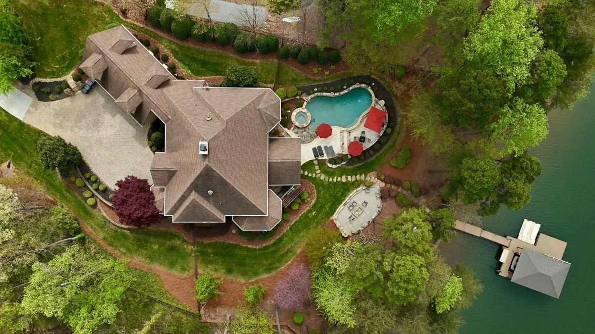 Aerial view of a house with a backyard pool, patio furniture, a dock on the water, surrounded by trees and landscaped gardens.