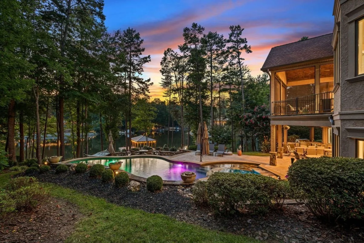 A backyard with a swimming pool, surrounded by trees and landscaped bushes. There are lounge chairs and umbrellas near the pool, with a house featuring a covered patio and balcony on the right. Specializing in commercial and lake front properties.