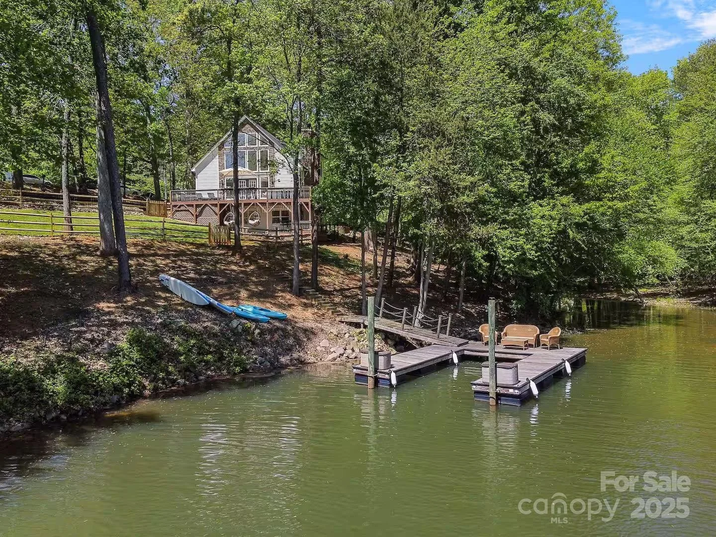 Lake with a wooden dock and seating area, house on hillside surrounded by trees, kayak on shore, and a lush green landscape under a clear blue sky.