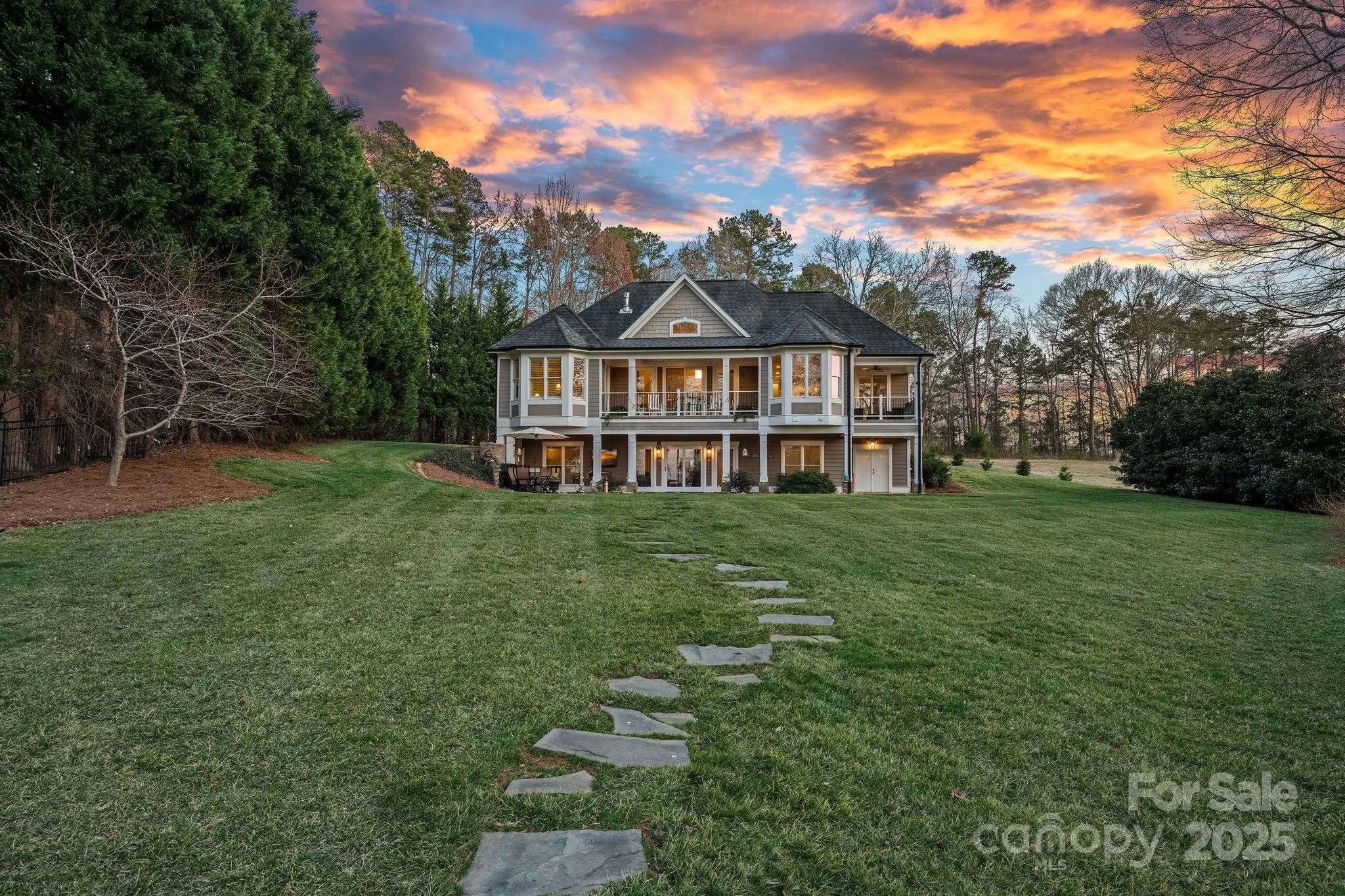 A large two-story house with a spacious backyard, a stone pathway leading to the house, and a sunset sky with orange and purple clouds.