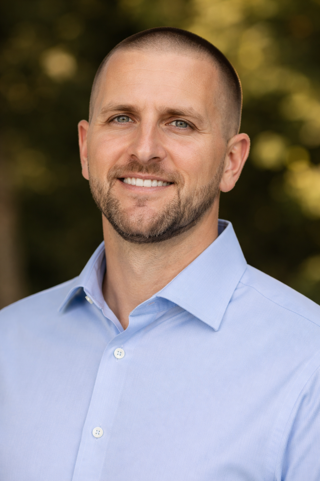 Portrait of a man with short hair and a beard, smiling, wearing a light blue dress shirt outdoors with blurred greenery in the background.