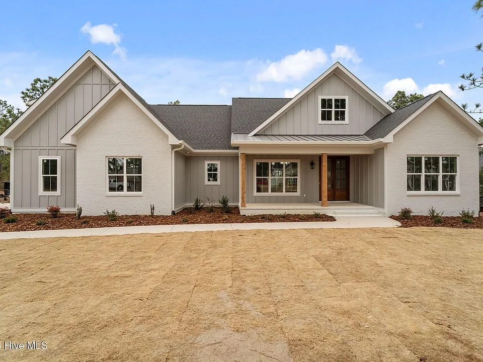 Newly constructed modern house with a combination of light gray siding and white brick, front porch with wooden support beams, multiple windows, and a clear blue sky.